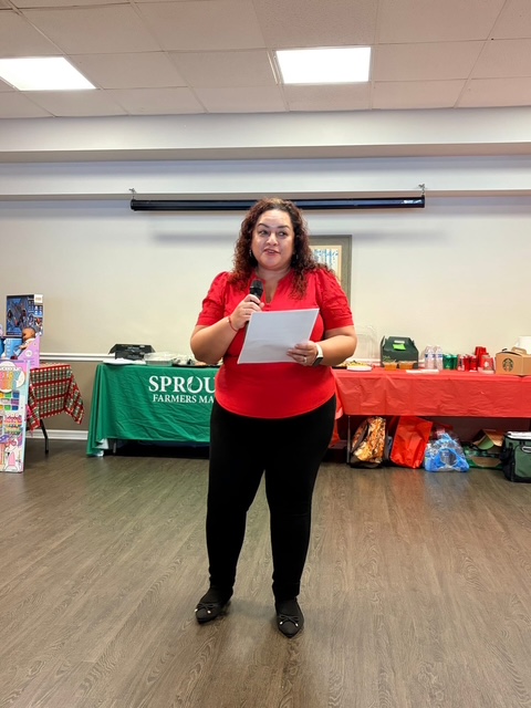 Woman with curly hair and glasses, wearing a red shirt, holding a paper, standing in a room with tables and a green banner.