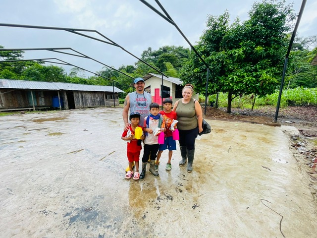 Group of five people standing outdoors on muddy ground with trees and a building in the background.