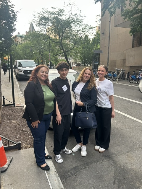 Four people standing on a sidewalk, smiling, with trees and buildings in the background.