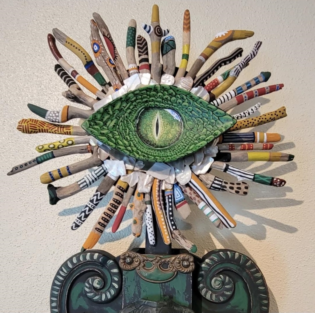 Mixed media artwork of a green eye with colorful, patterned paper strips radiating outward, mounted on a decorative base.
