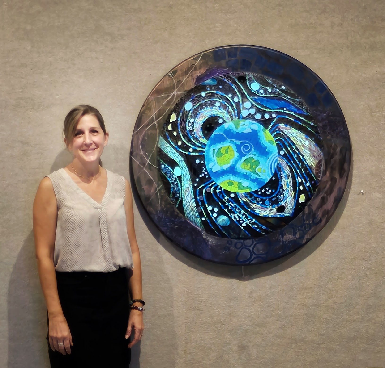 Woman standing next to a circular artwork with swirling blue and green patterns on a gray wall.