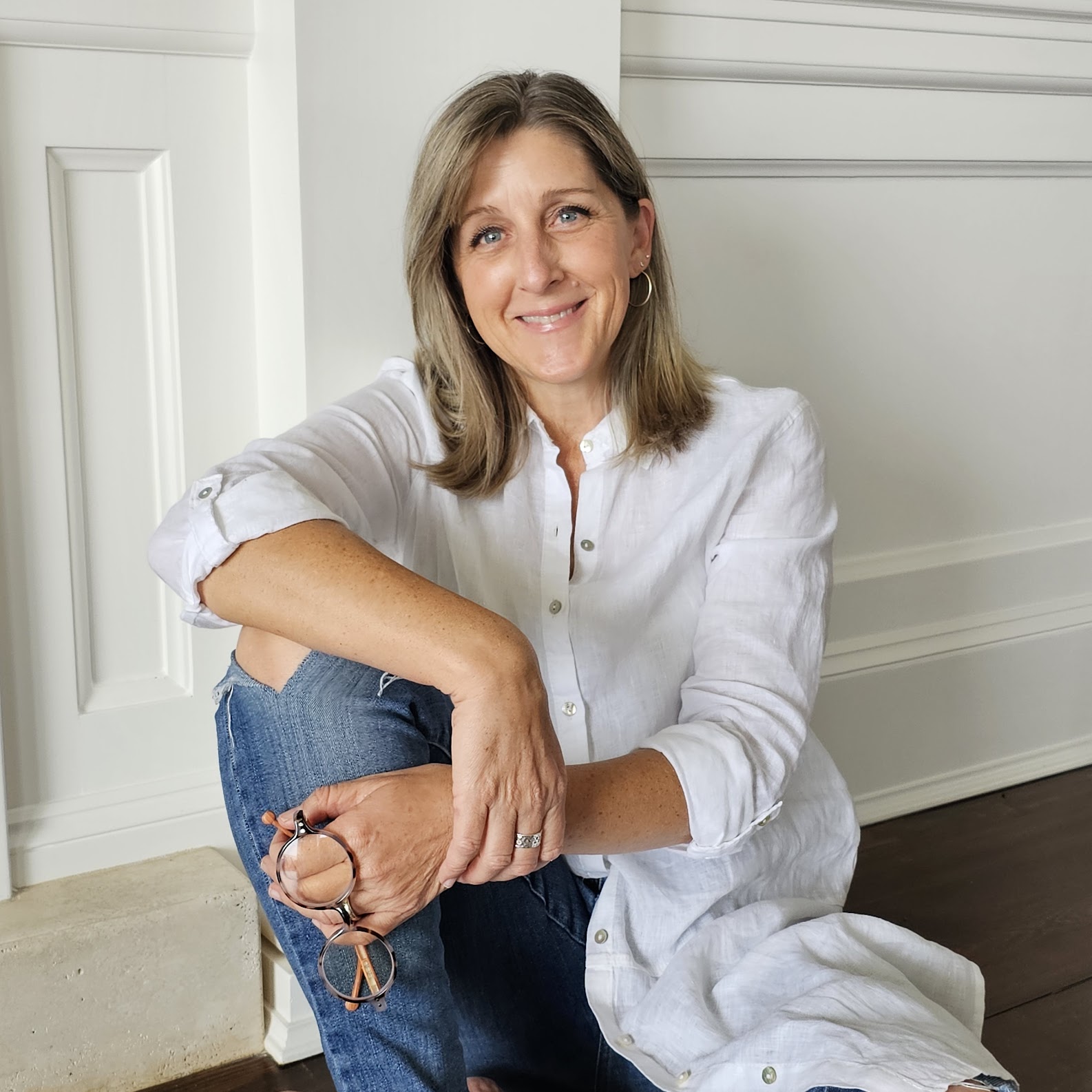 Woman with shoulder-length hair smiling, sitting on the floor with her arm resting on her knee, wearing a white shirt and jeans, in a bright room.