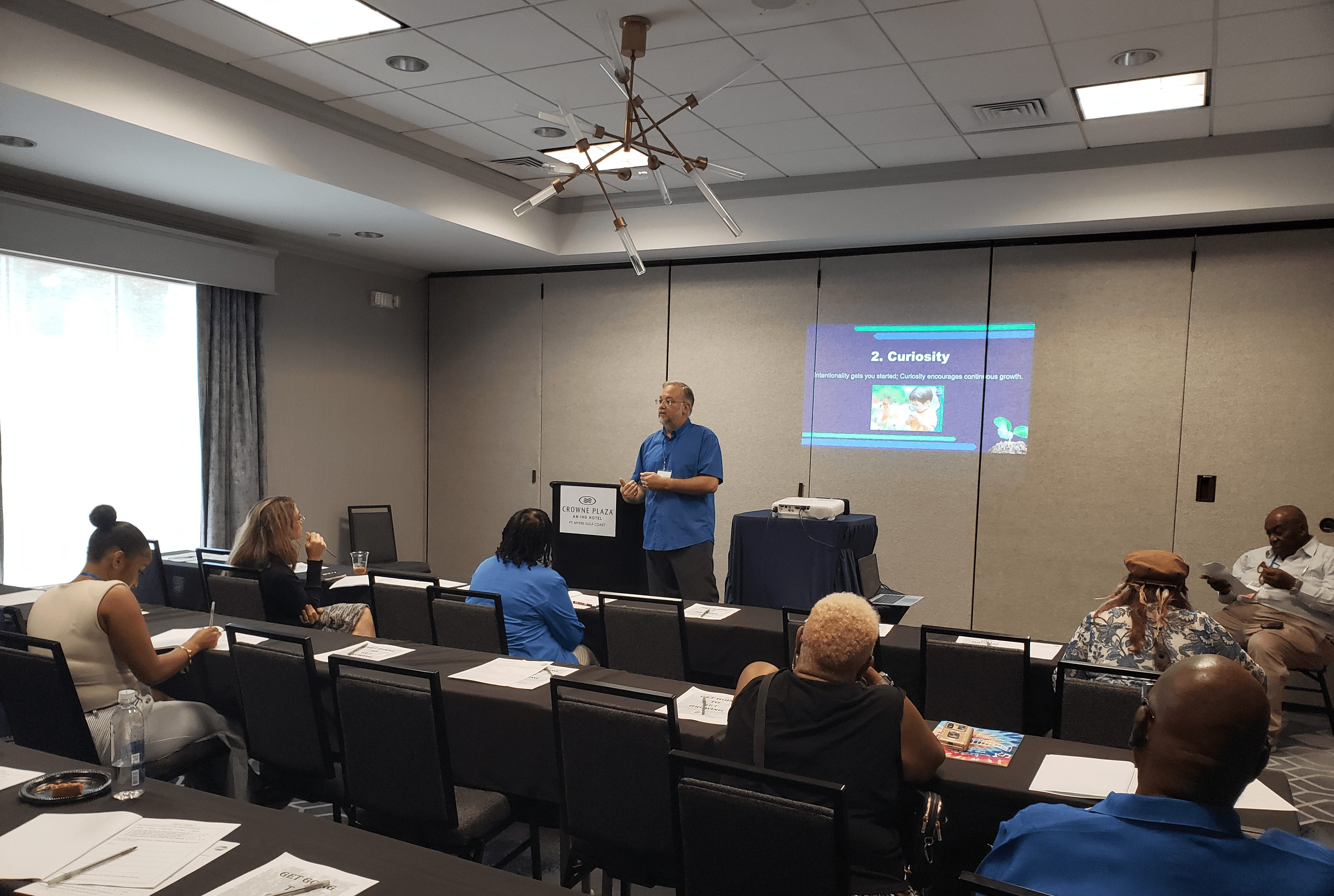 Person giving presentation in a conference room with seated audience, projector screen displaying slides, and a ceiling light fixture.