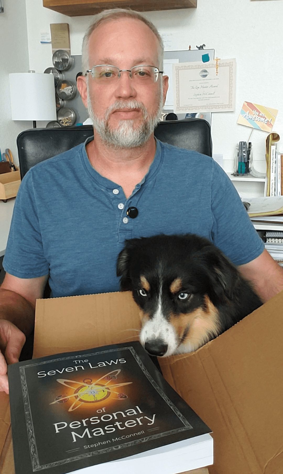 Man with glasses and beard sitting at desk with a dog in a box, book titled 'Seven Laws of Personal Mastery' visible.