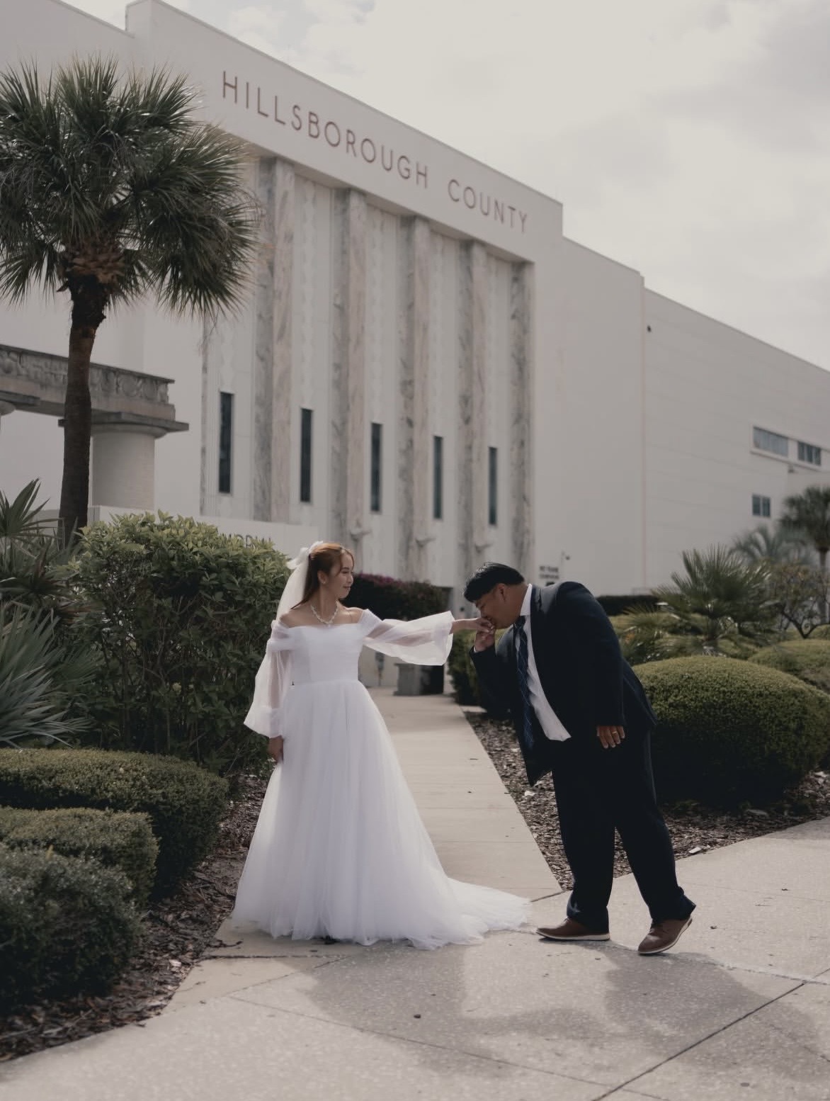 A bride in a white wedding dress and a groom in a dark suit are outside a building labeled 'Hillsborough County.' The bride is touching the groom's face, and they are smiling. There are bushes and a palm tree nearby.