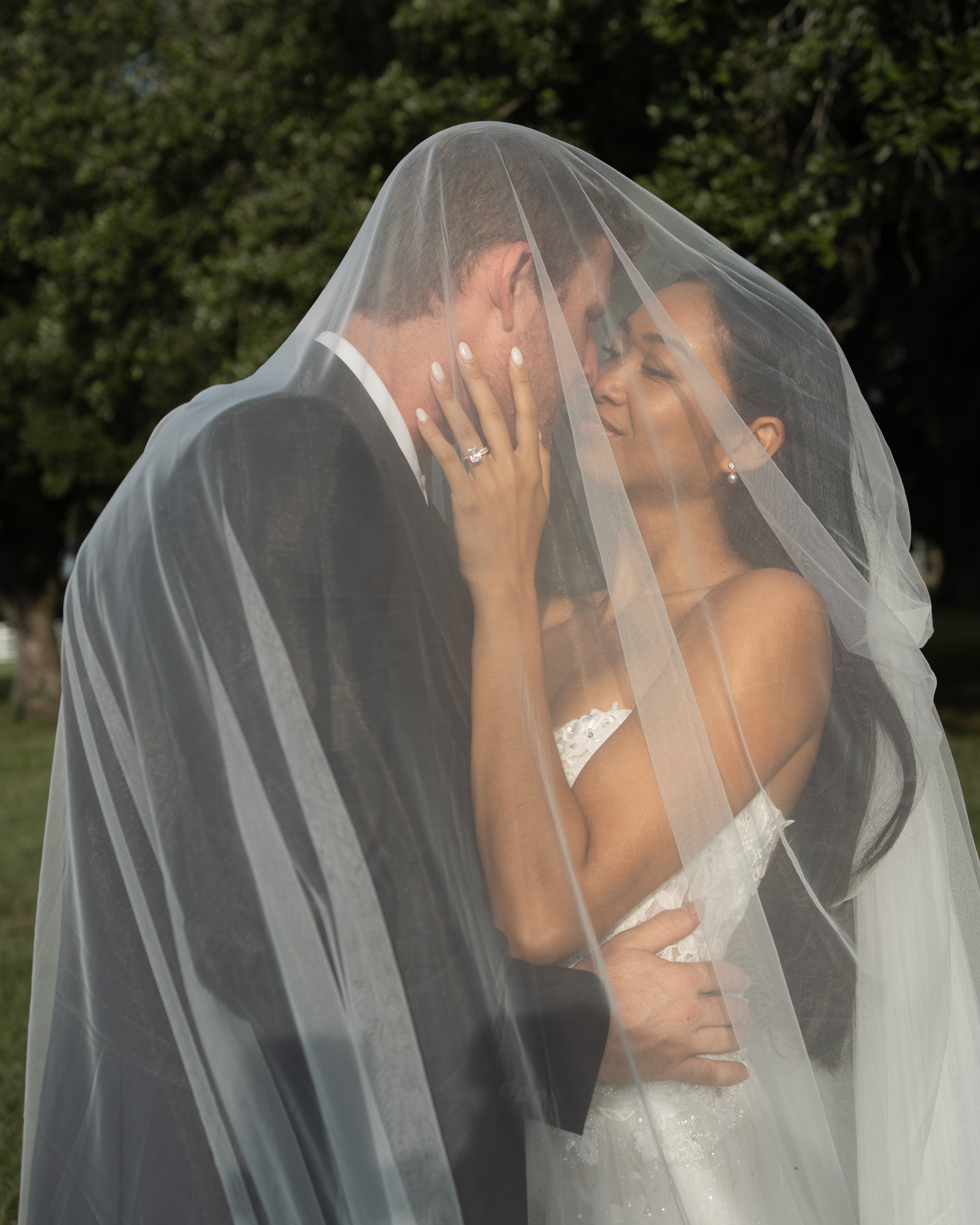 Bride and groom share a kiss under a veil outdoors, with trees in the background.