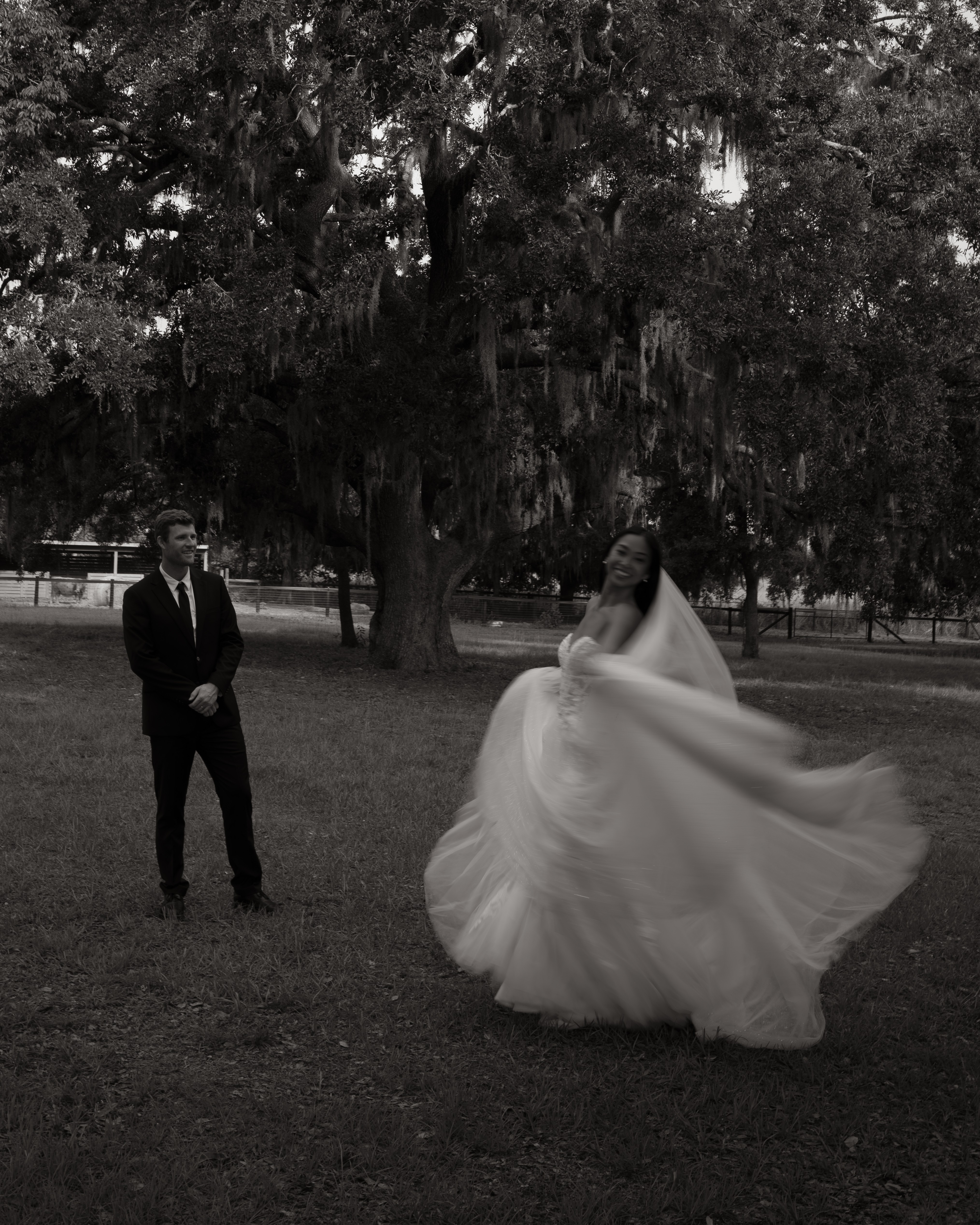A bride in a flowing wedding dress twirls outdoors with a groom nearby, under large trees in a park.