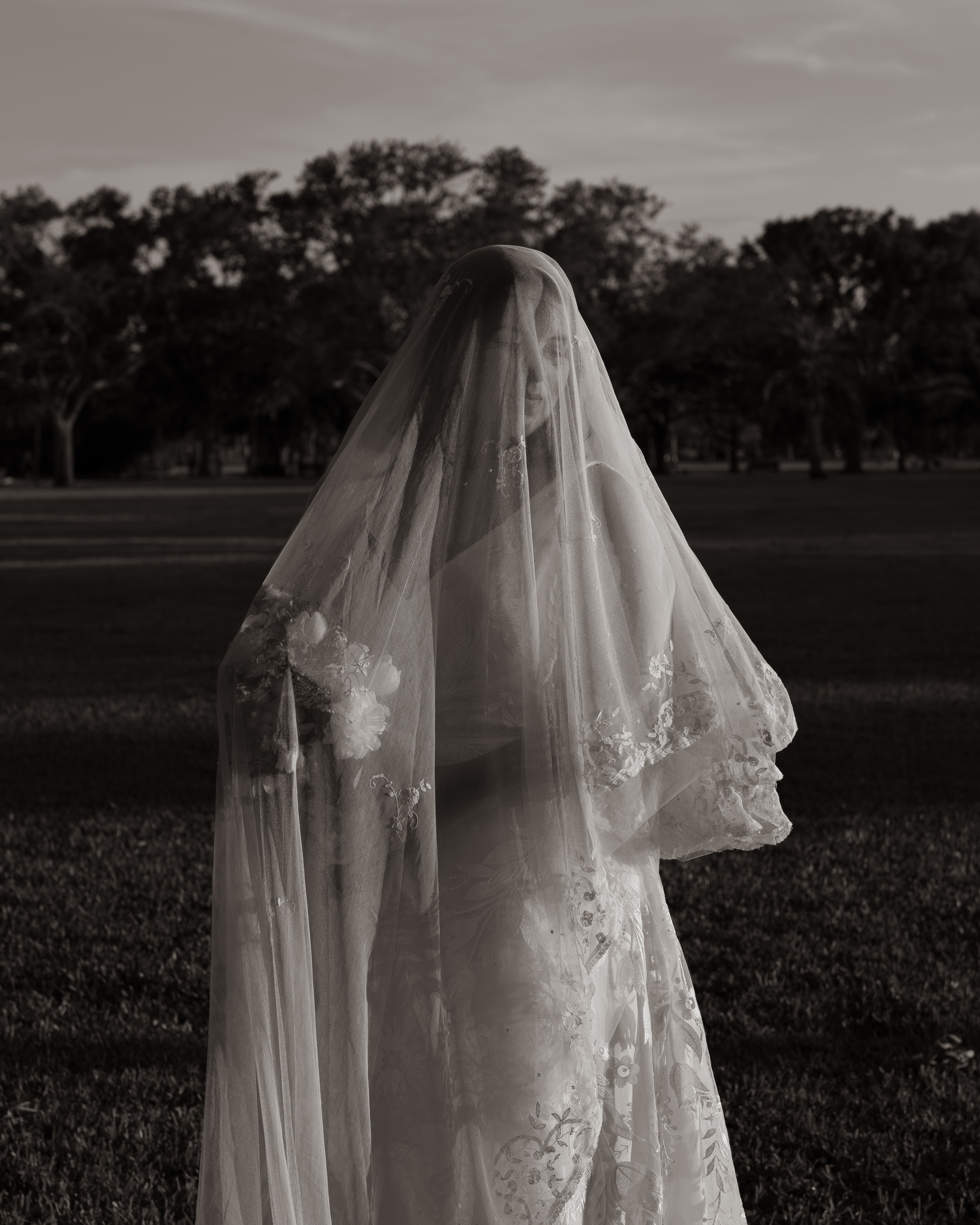 Person wearing a veil outdoors with trees in the background, facing away, in black and white.