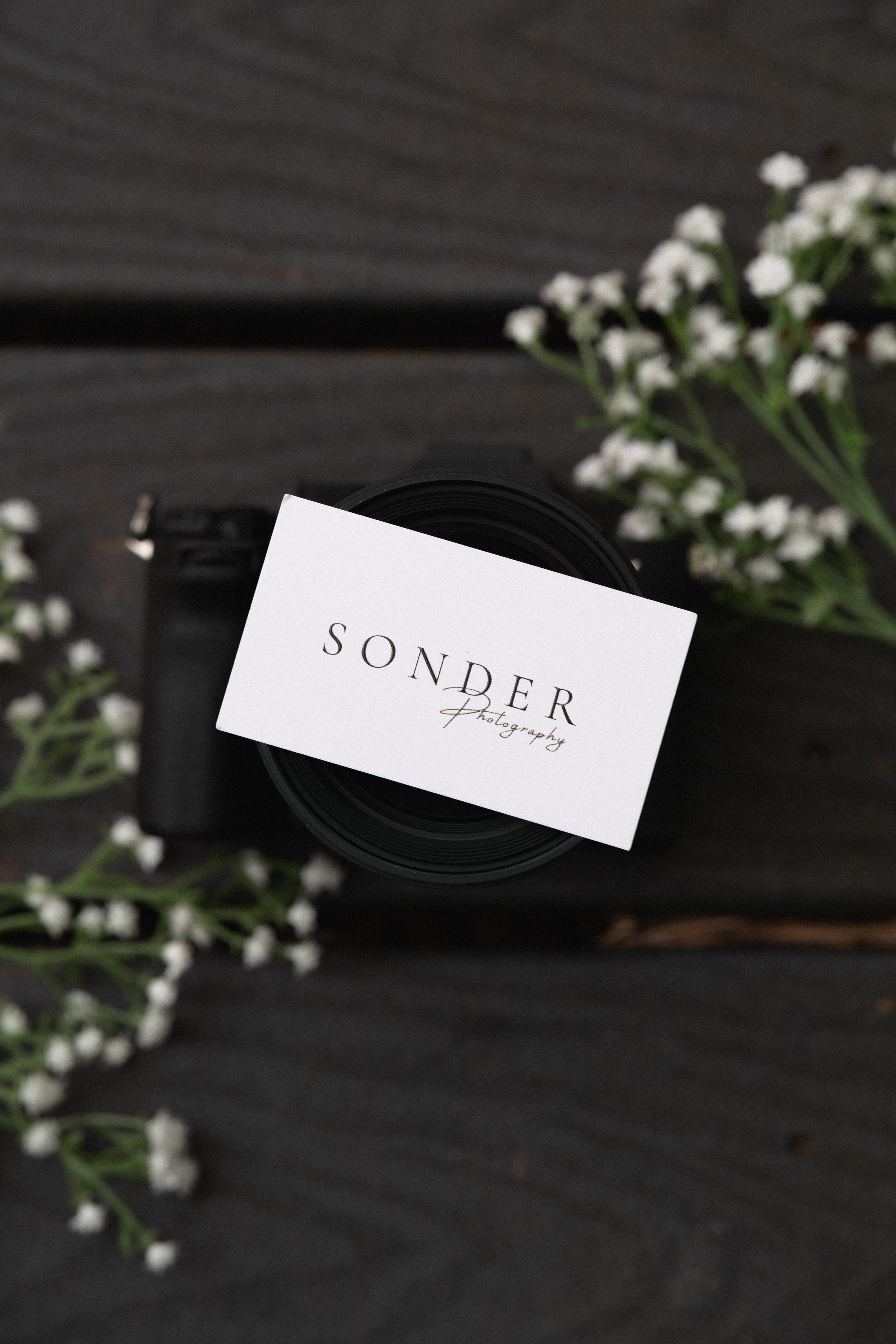 Camera lens with a business card reading 'SONDER photography' on top, surrounded by white flowers on a dark wooden surface.