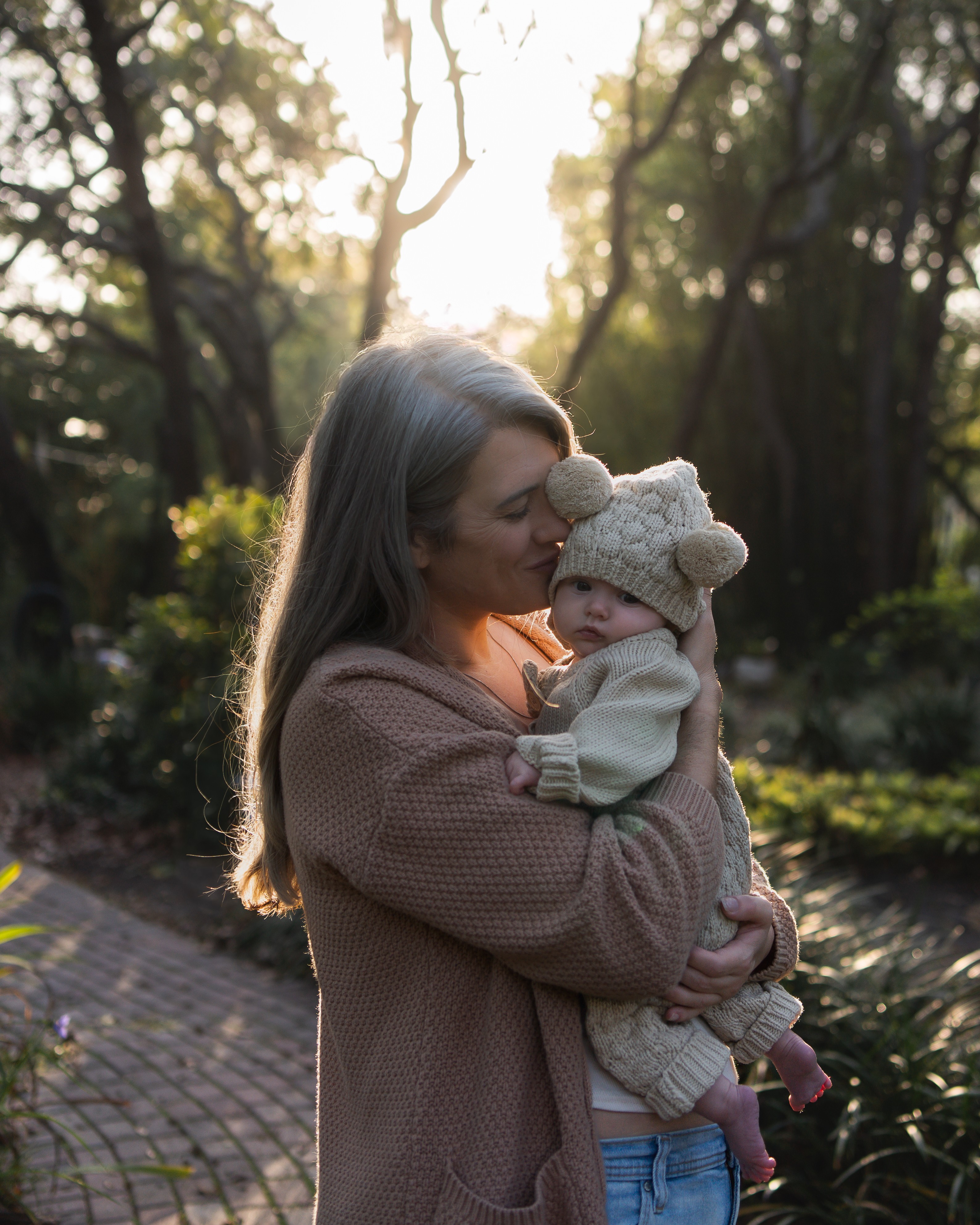 Woman holding a baby outdoors in a wooded area during sunlight, woman wearing a brown sweater, baby in a knit hat and sweater.