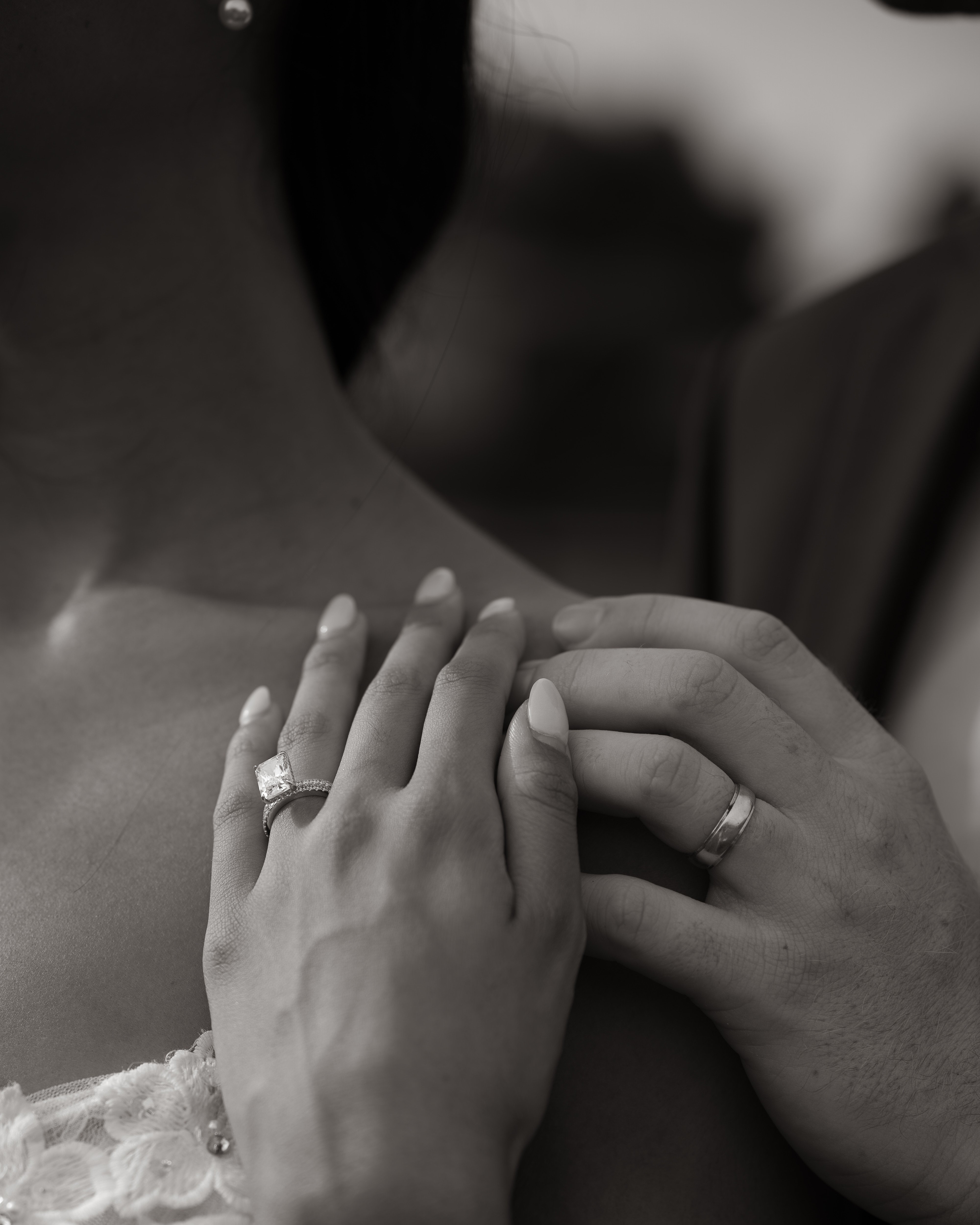 Two people hold hands, showing rings on their fingers, with a blurred background, in black and white.