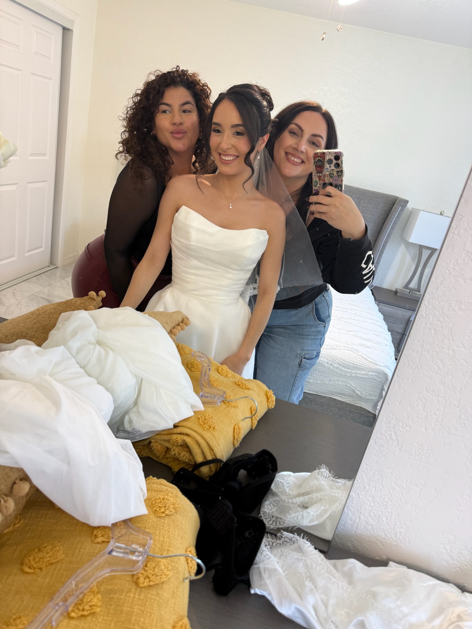 Three women taking a mirror selfie in a bedroom, smiling, with a bed and clothes in the foreground.