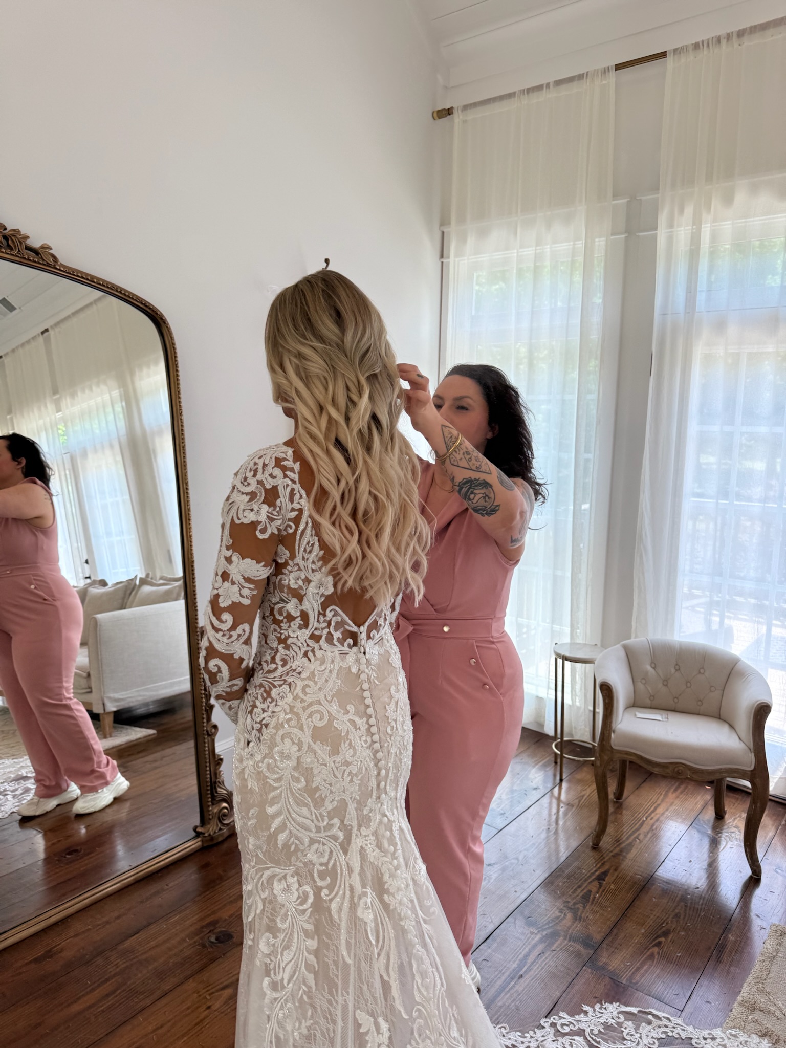 Woman in a lace wedding dress is assisted by another woman in a pink jumpsuit, in a bright room with a mirror and armchair.