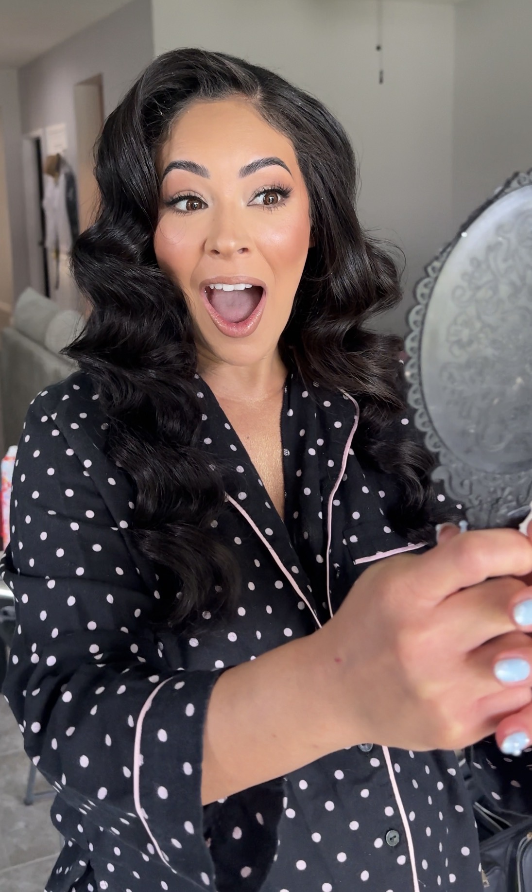 Woman with long dark hair in polka dot pajamas looks surprised while holding a mirror.
