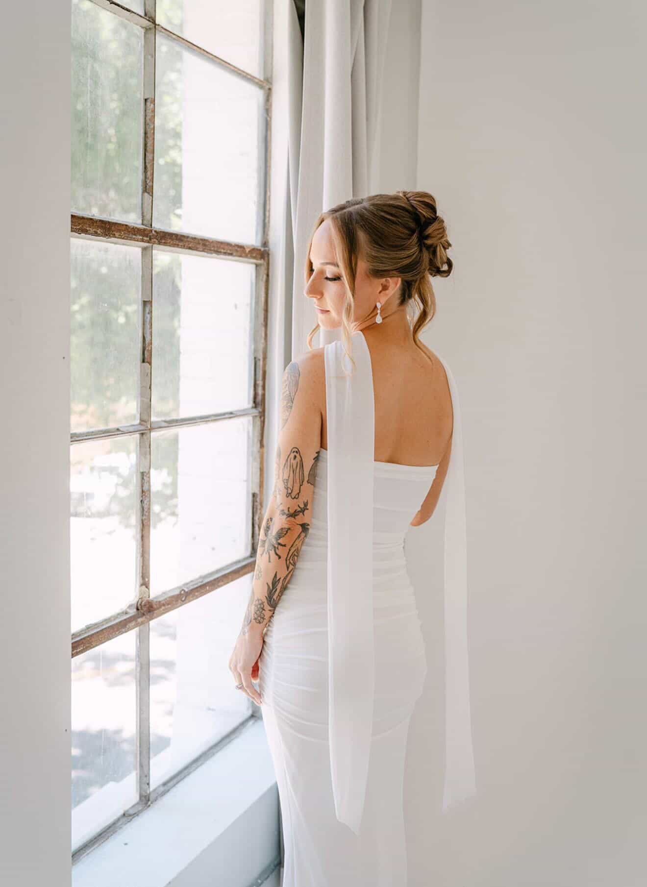 Woman in white dress with tattoos looking out window, natural light, indoor setting, hair in bun, side profile.