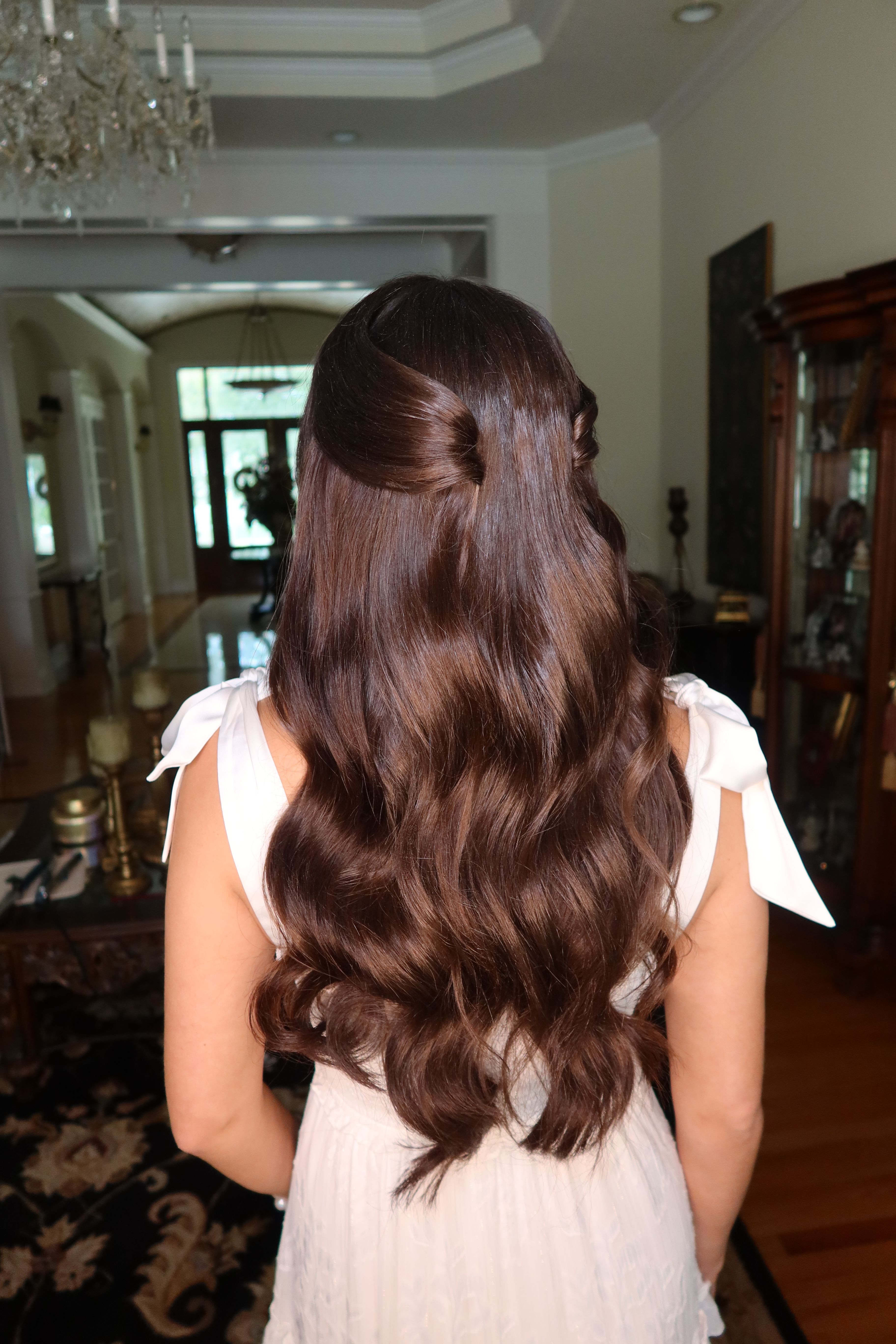 Person with long, wavy brown hair wearing a light-colored dress, standing indoors with furniture and a window in the background.