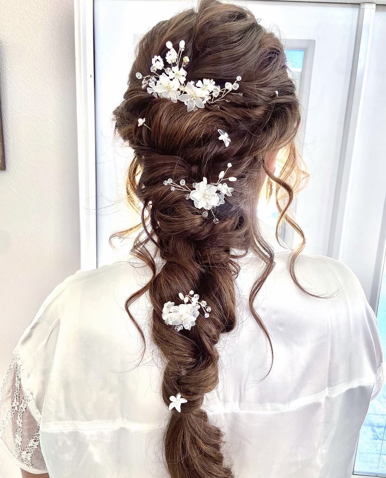 Brunette hairstyle with floral hair accessories and loose curls, viewed from the back, on a person wearing a white garment.
