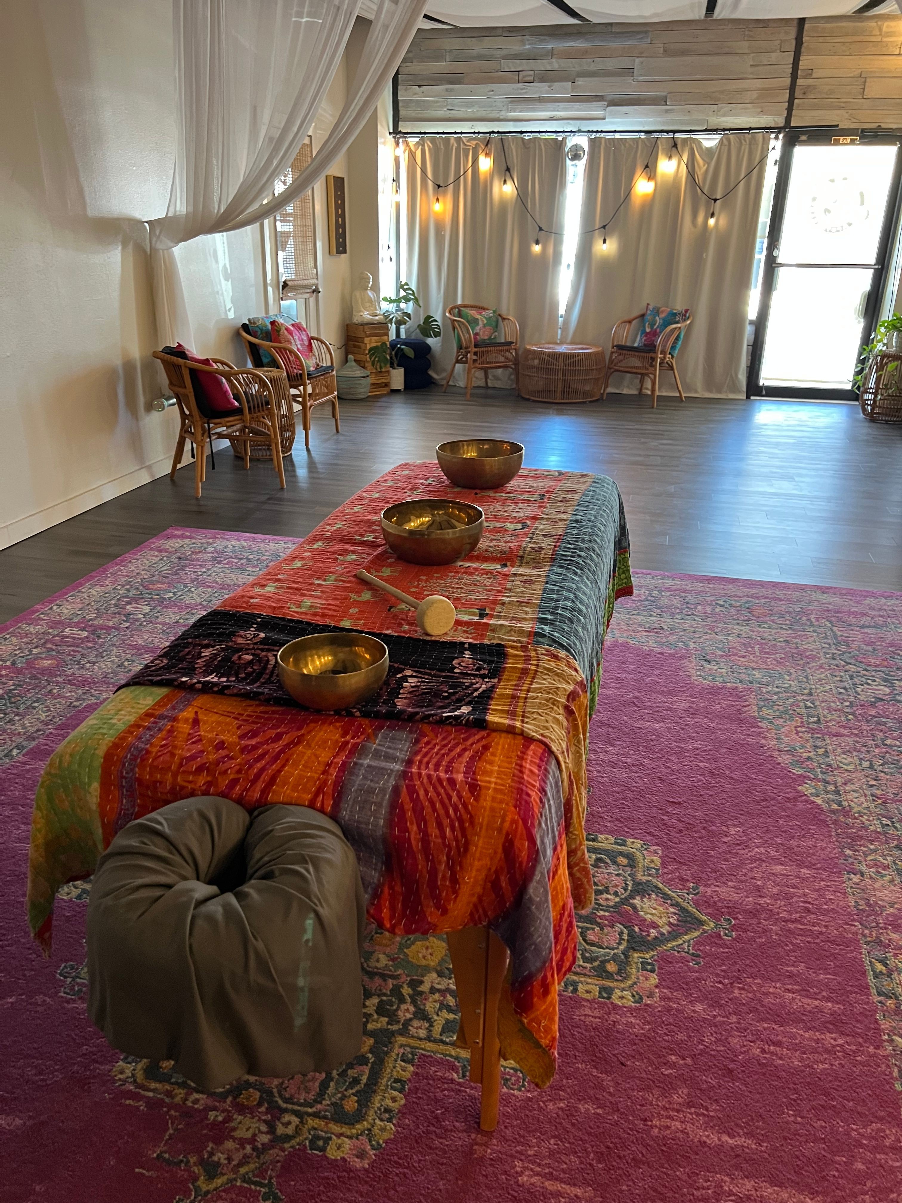 Person sitting on a colorful, patterned rug with a low table and bowls, in a room with large windows and wooden floors.