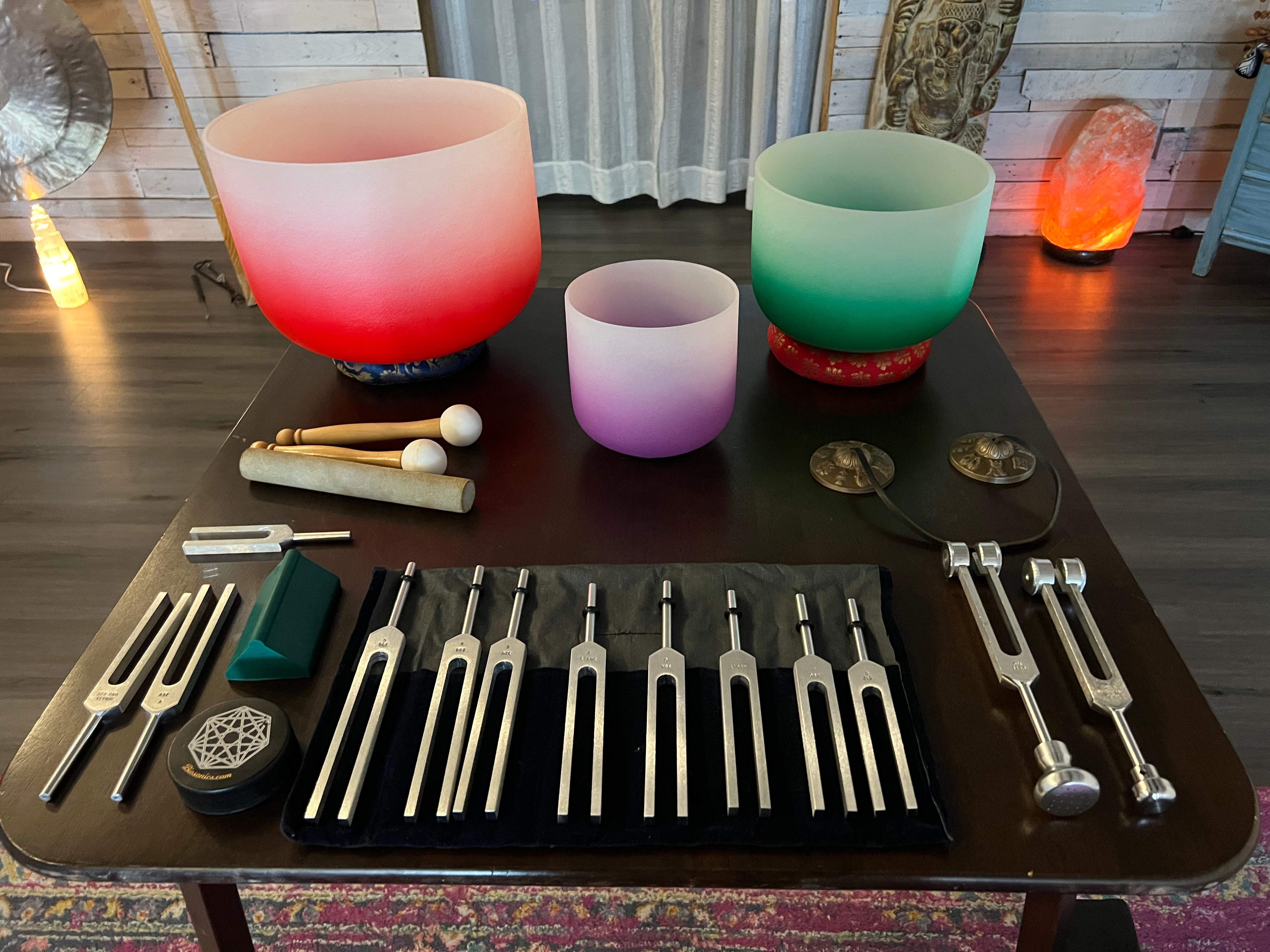 Table with colorful singing bowls, mallets, and tuning forks, set on a dark wooden surface in a room.