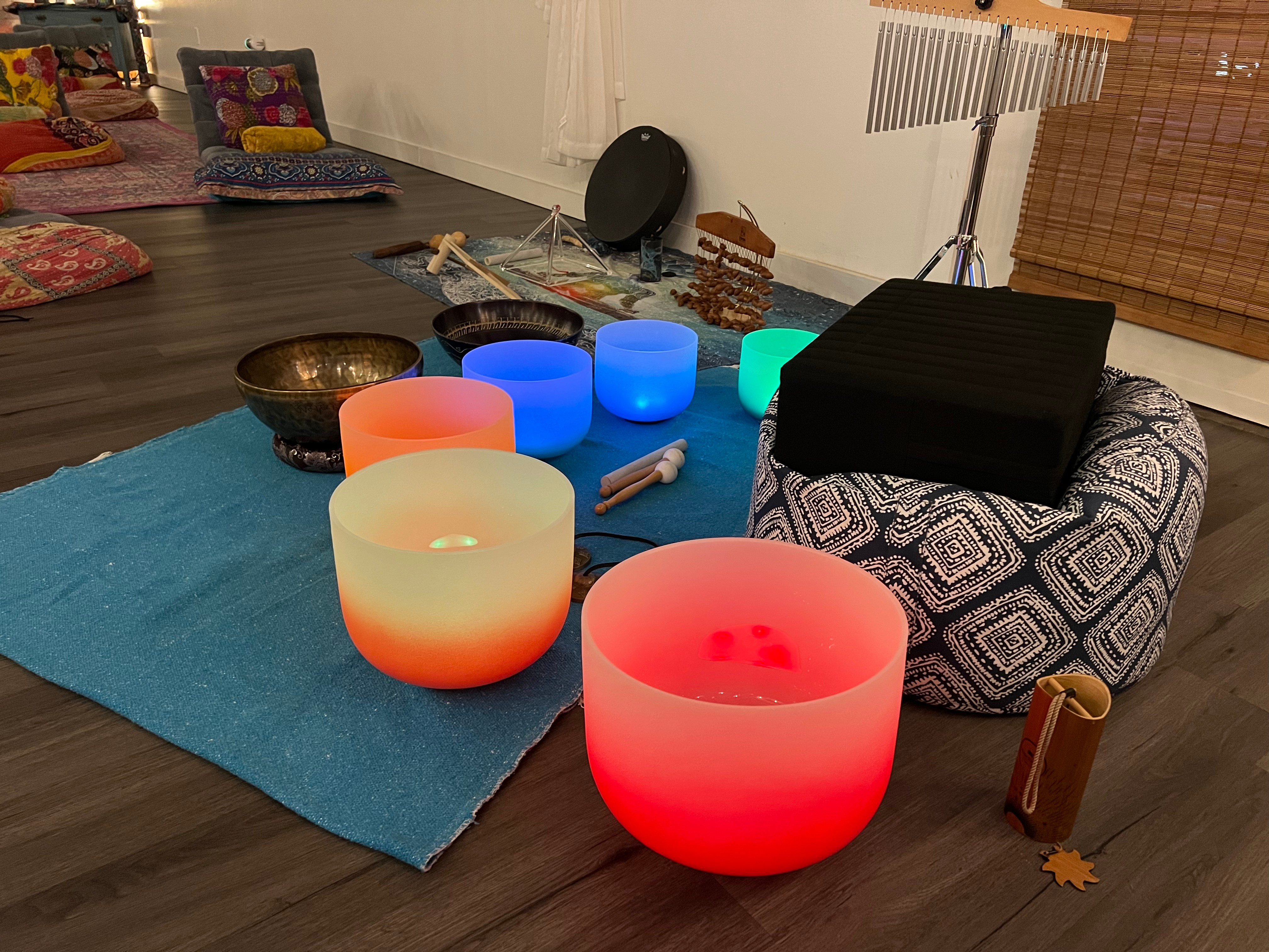 Room with colorful singing bowls, cushions, and a blue rug on dark wood floor, with a black box and patterned cushion nearby.