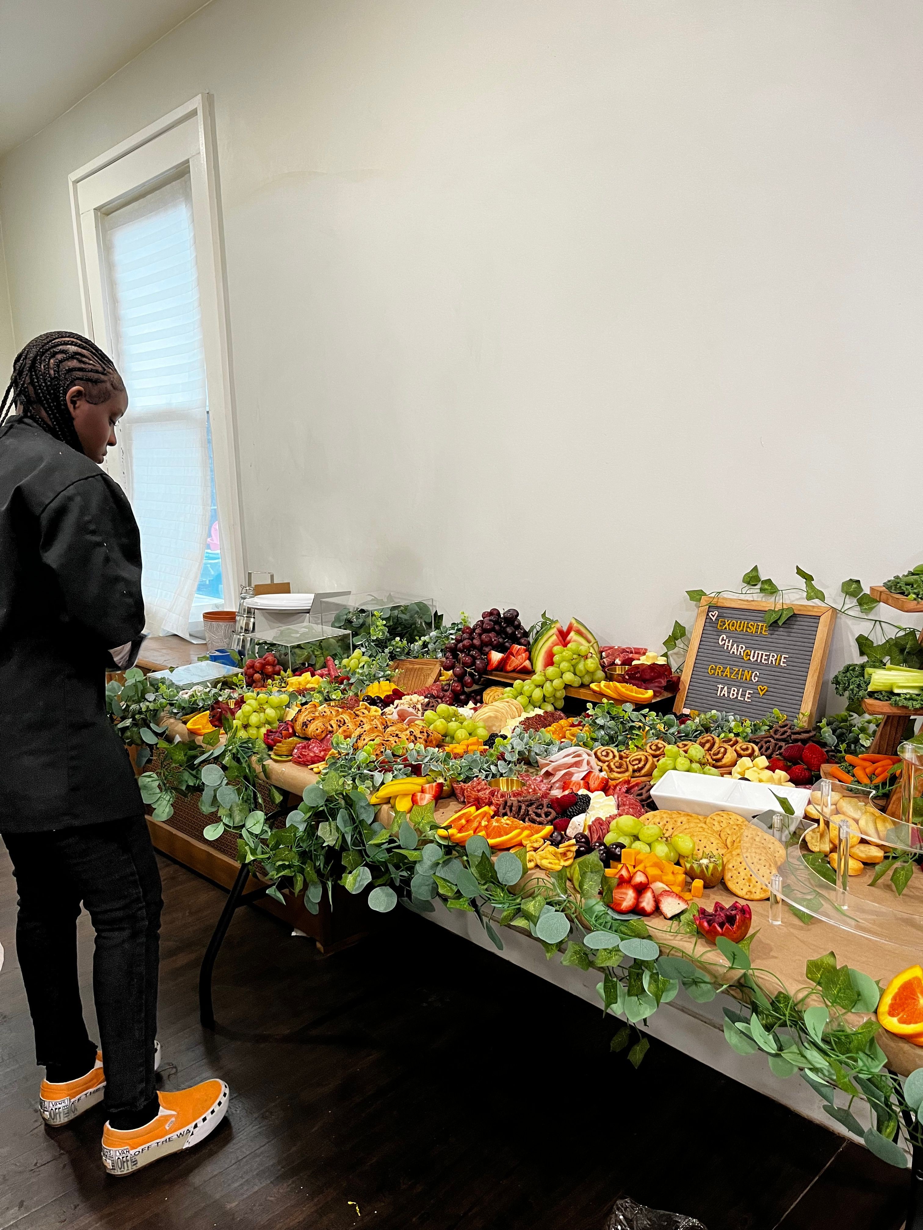 Person standing next to a table with a colorful fruit and vegetable display, in a room with a window and white wall.