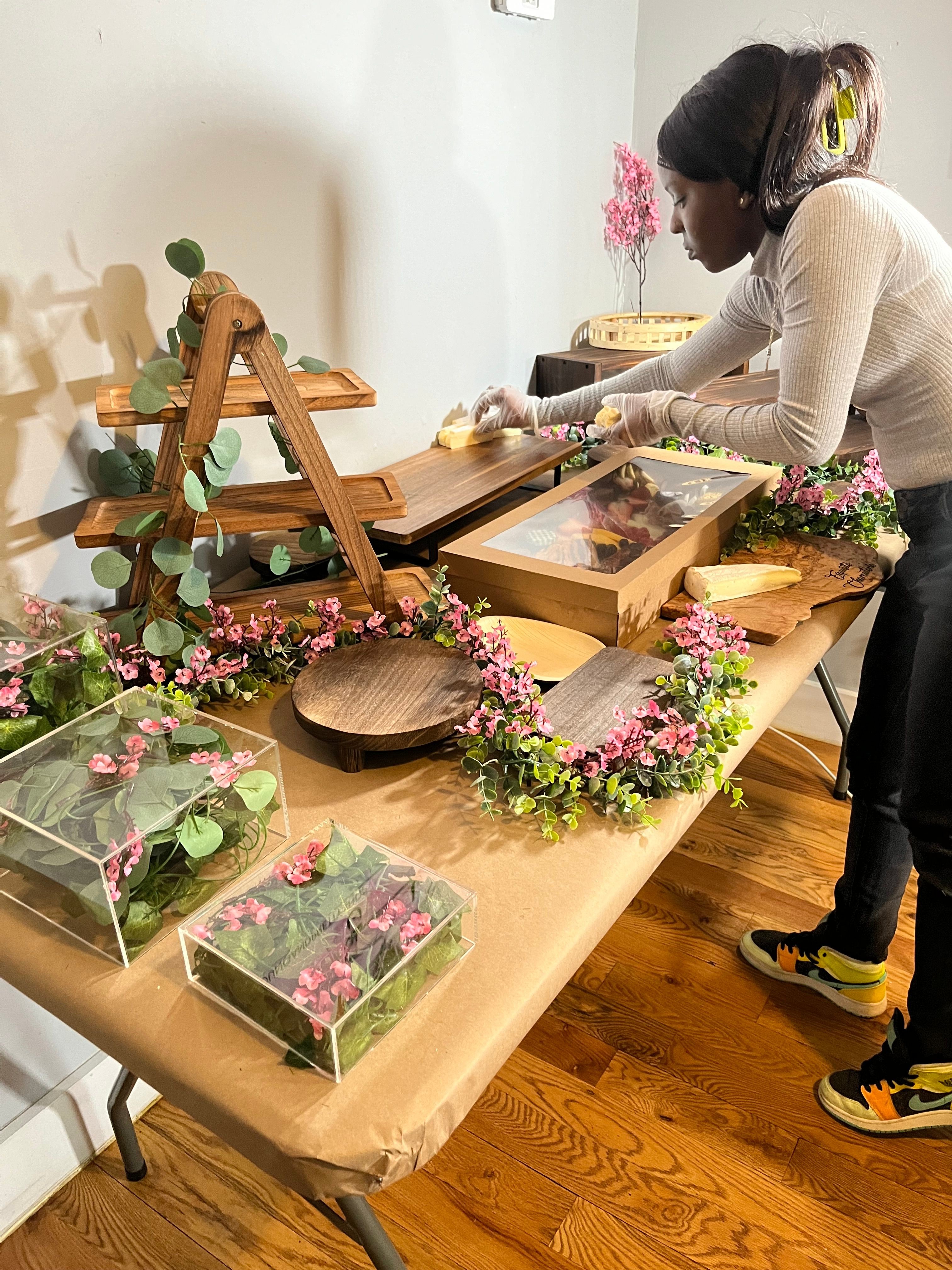 Person arranging flowers on a table with floral decorations and framed pictures, indoors with wooden flooring.