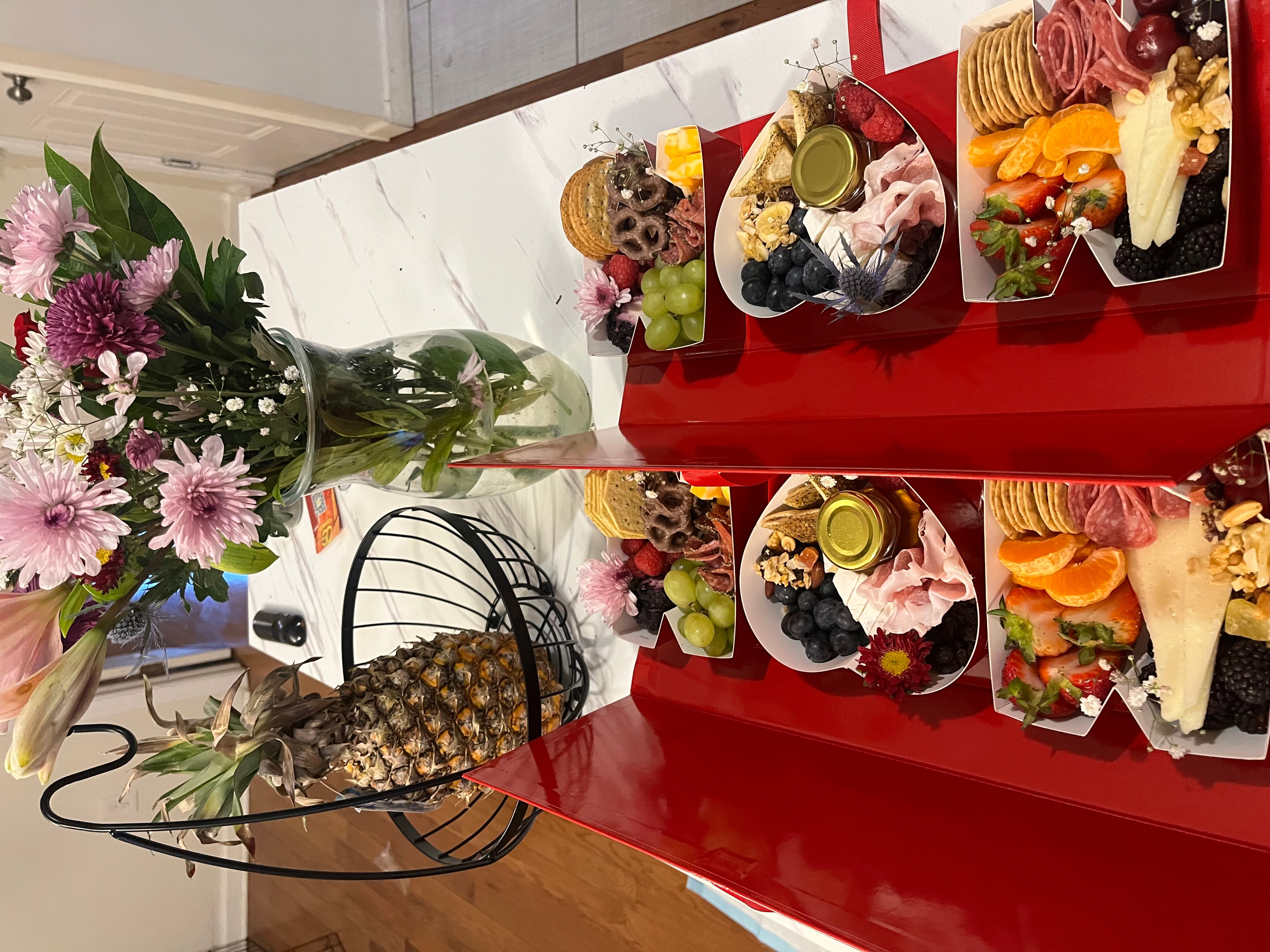 Table with fruit, desserts, and flowers, viewed from the side, with a red surface and a bouquet of flowers.