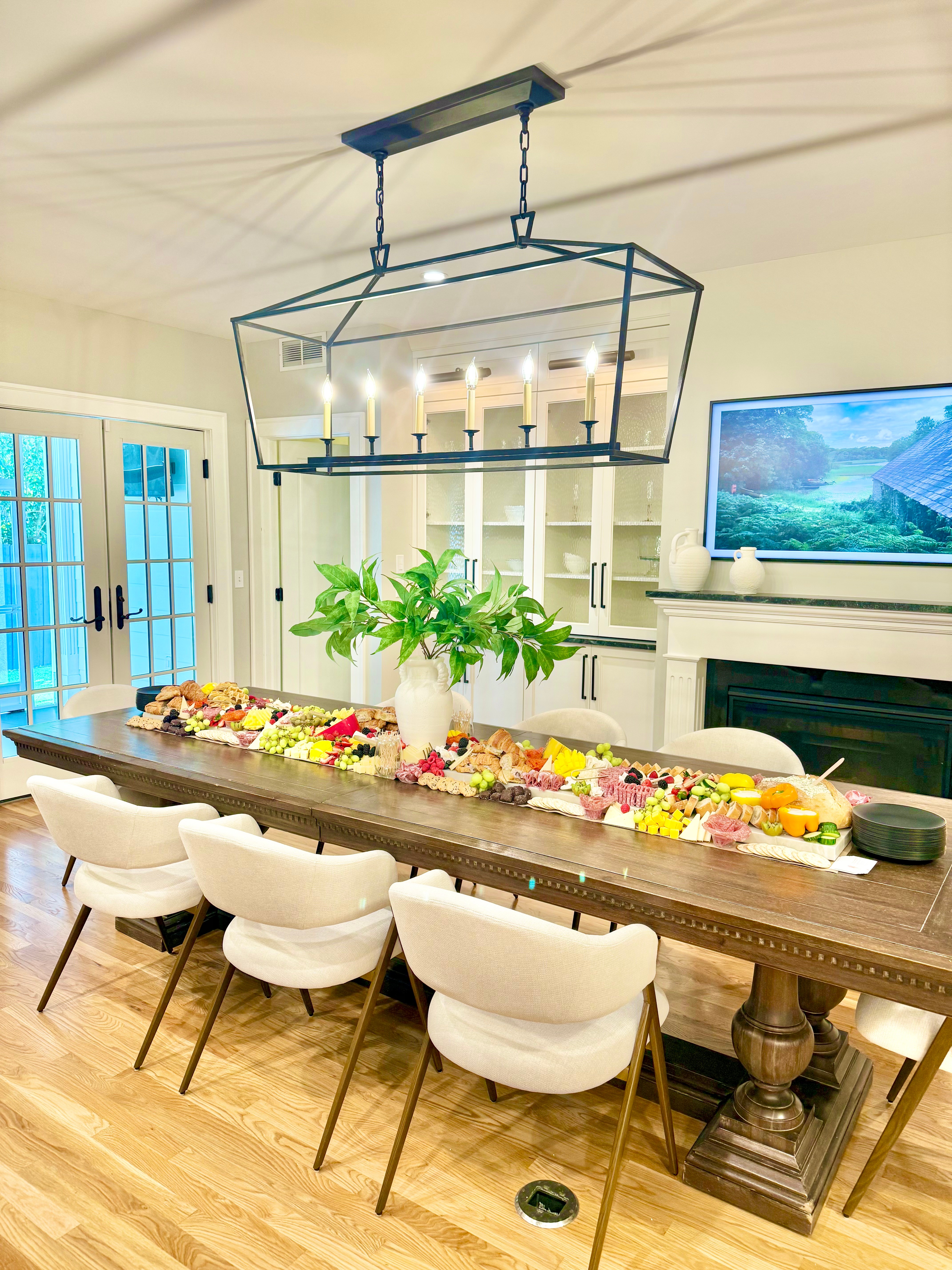 Dining room with a long table, white chairs, a chandelier, a fireplace, and a TV on the wall, decorated for a celebration.
