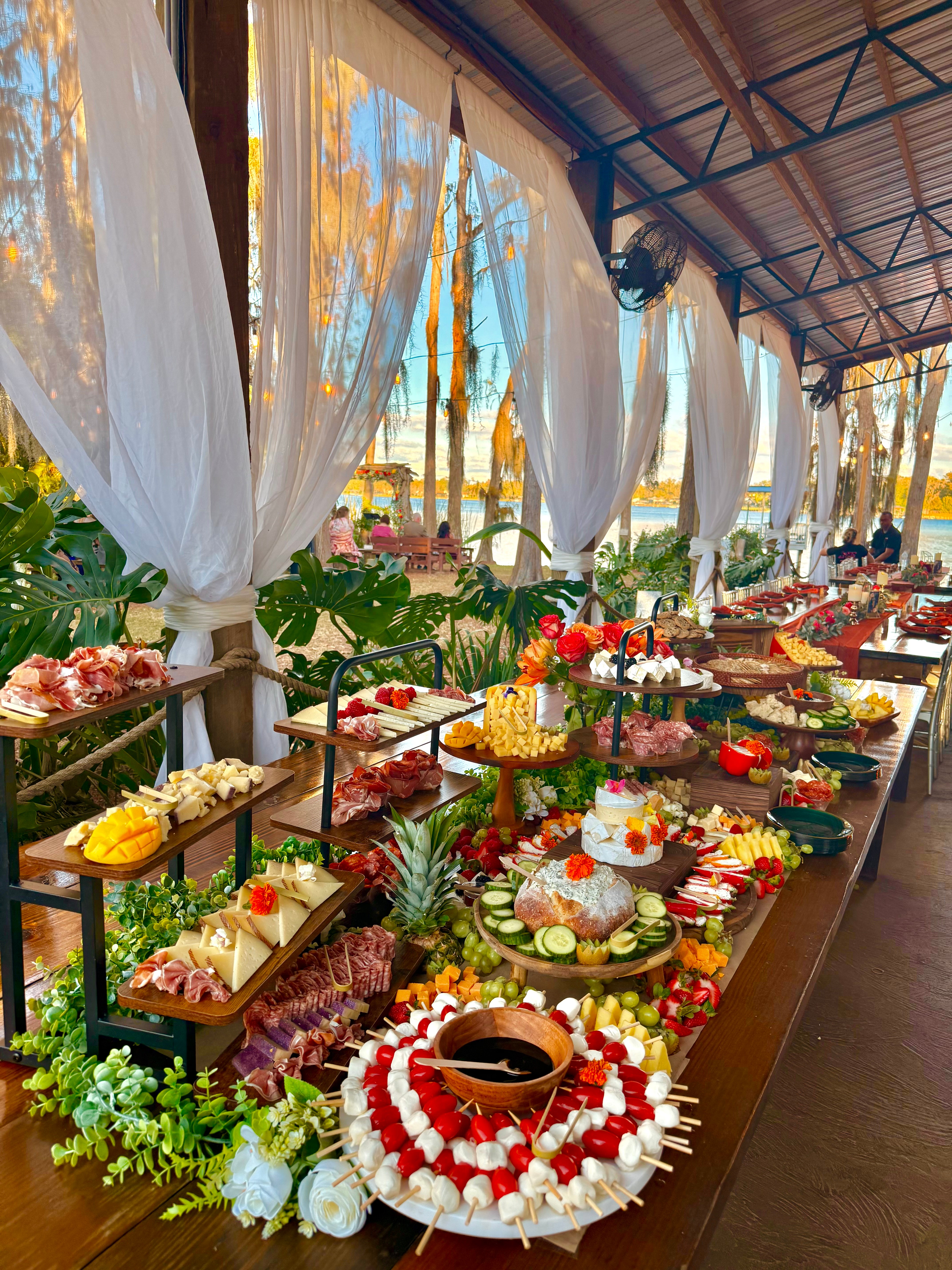 Long table with decorated desserts and flowers under a canopy with white curtains, outdoor setting with trees and sky visible.