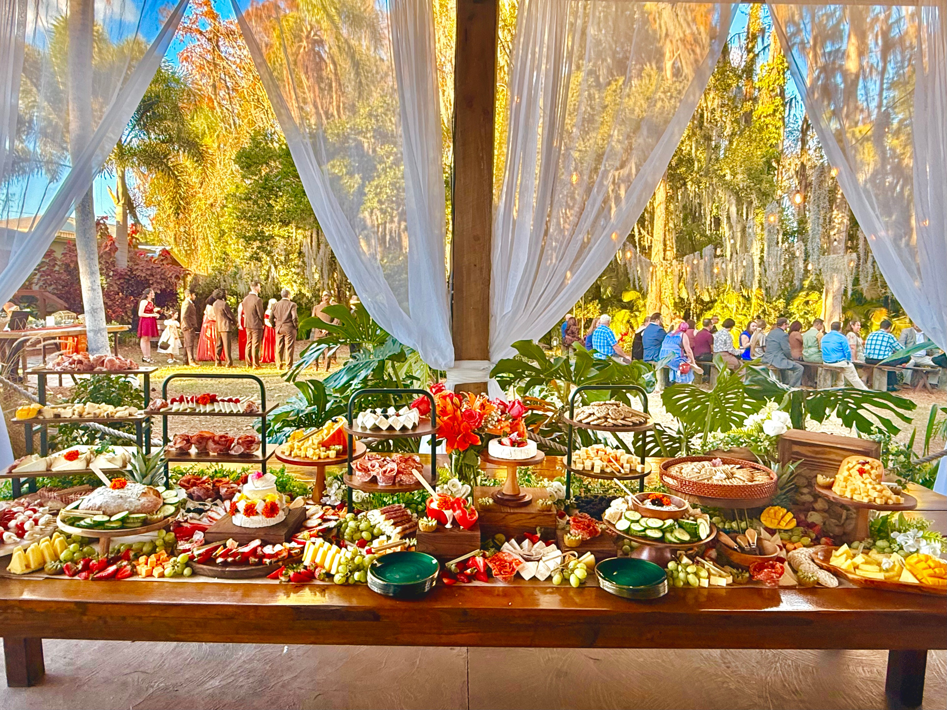 Dessert table with cakes, fruits, and decorations outdoors, with trees and people in the background.