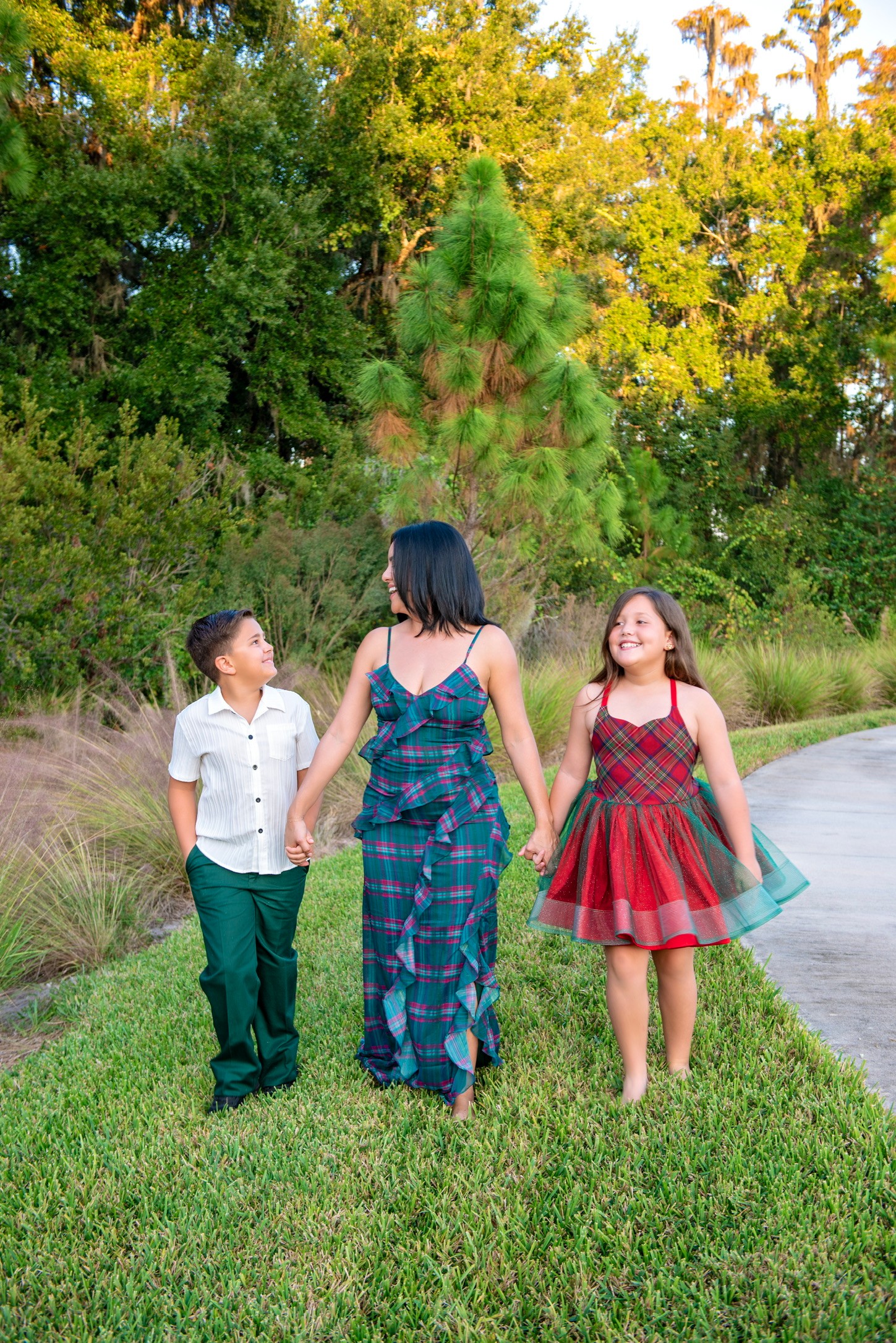 Woman walking with two children on grass near a pond, trees in background, sunny day.