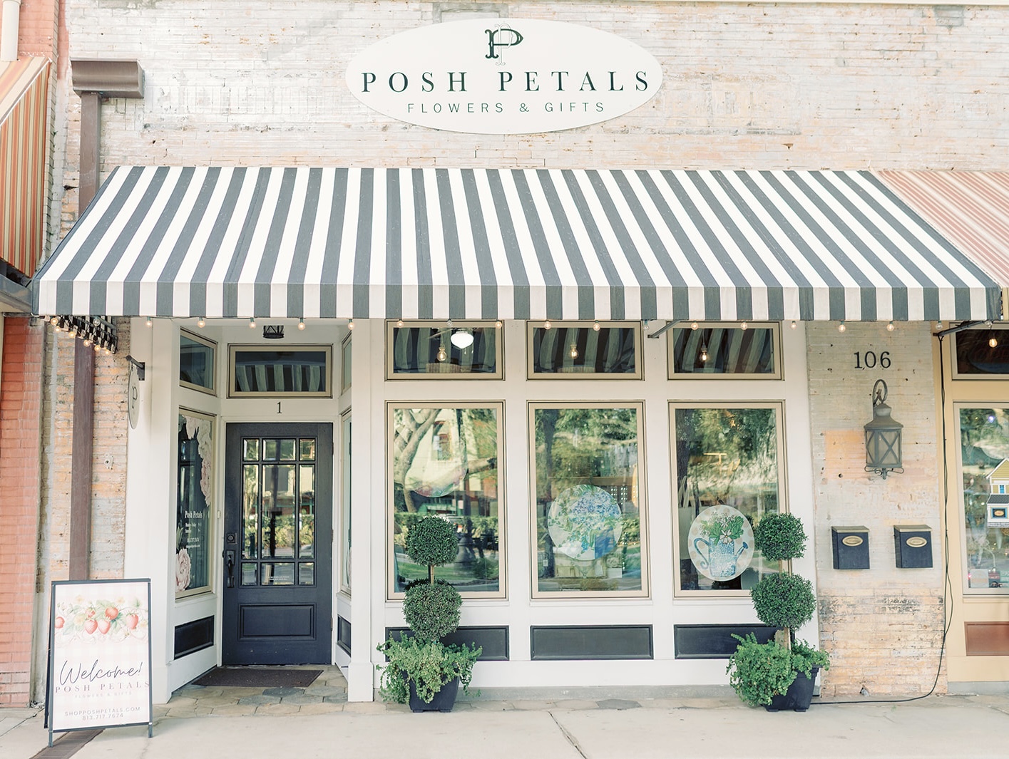 Storefront with striped awning, potted plants, and a sign reading 'Posh Petals Flowers & Gifts'.