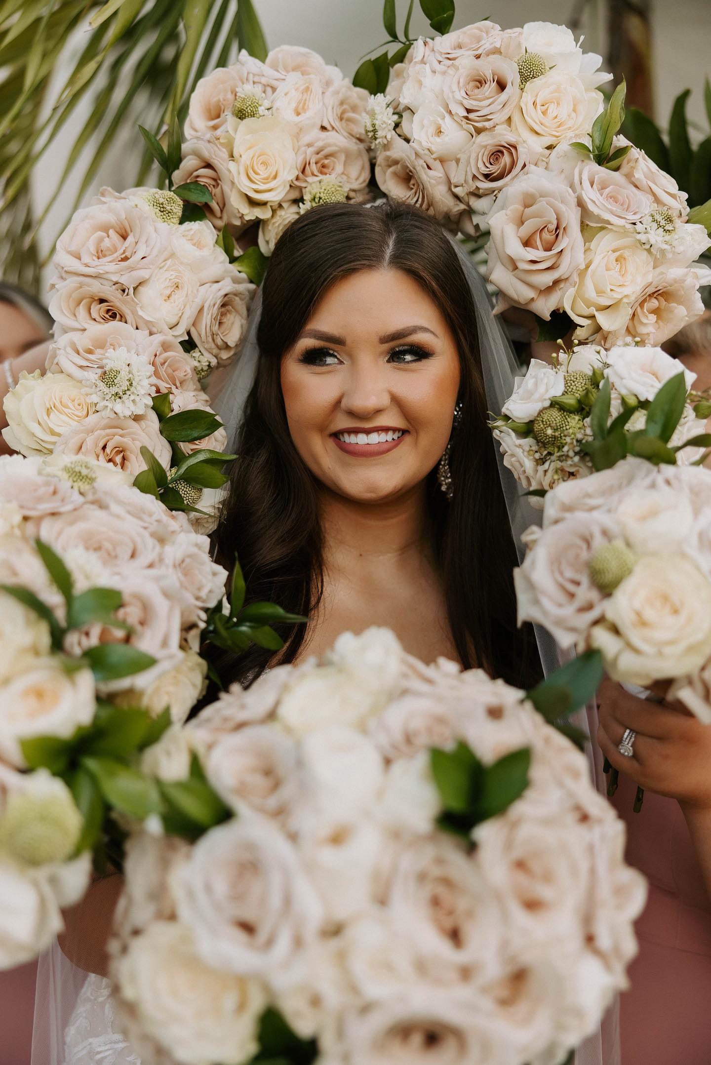 Woman with dark hair smiling surrounded by large cream and blush roses, floral arrangement forming a circle around her face.