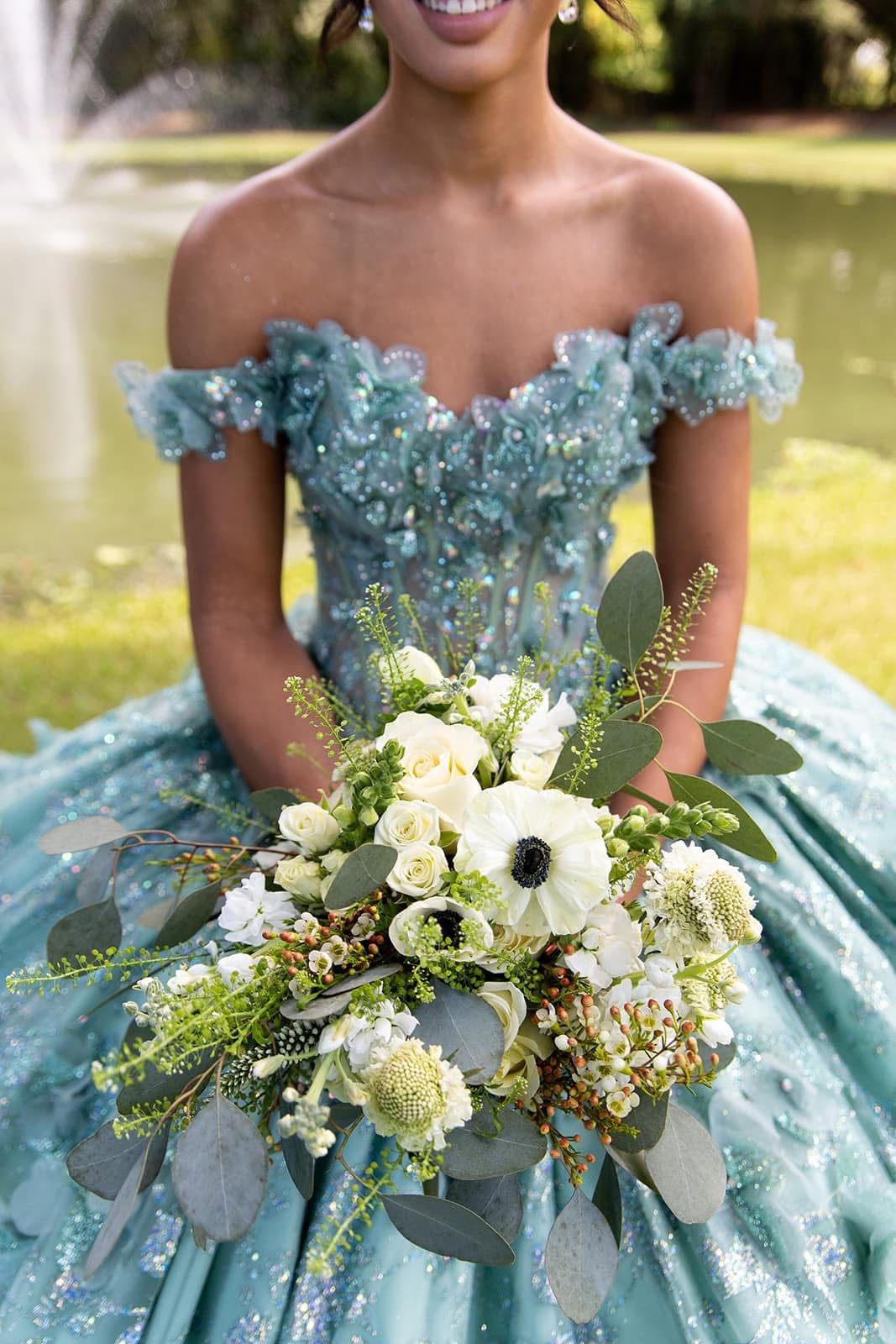 Woman in a blue dress holding a bouquet of white and green flowers outdoors, smiling, with a lake in the background.