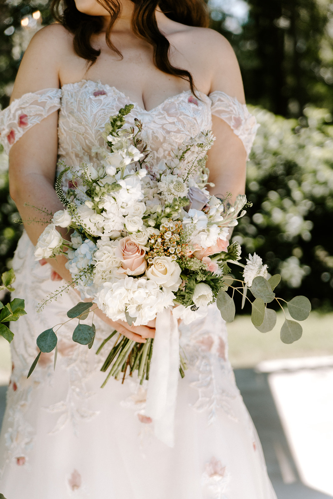 Person in wedding dress holding a bouquet of white and pink flowers outdoors, with trees in background.