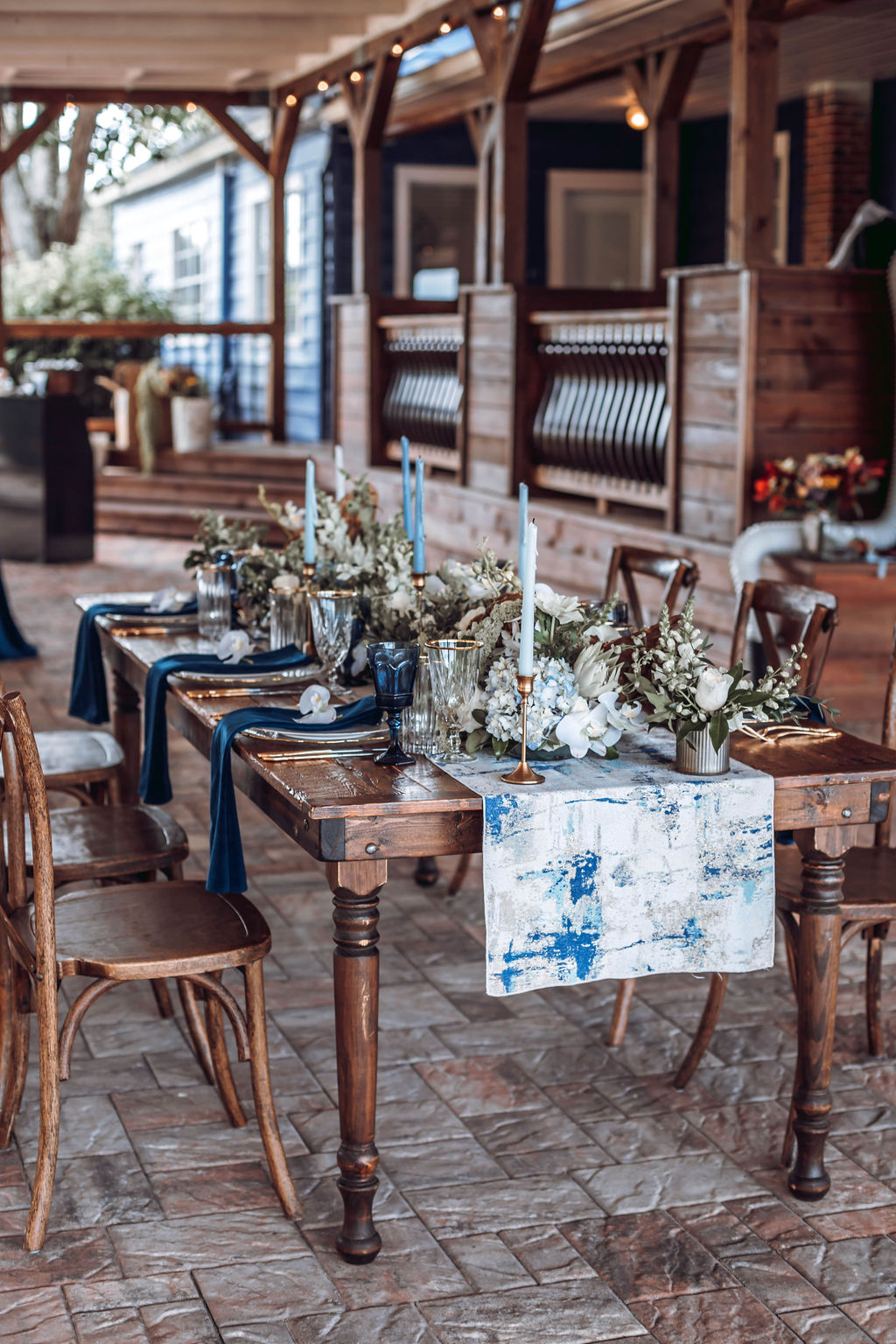 Outdoor dining table set with floral arrangements, chairs, and a table runner, on a stone patio with a wooden porch in background.