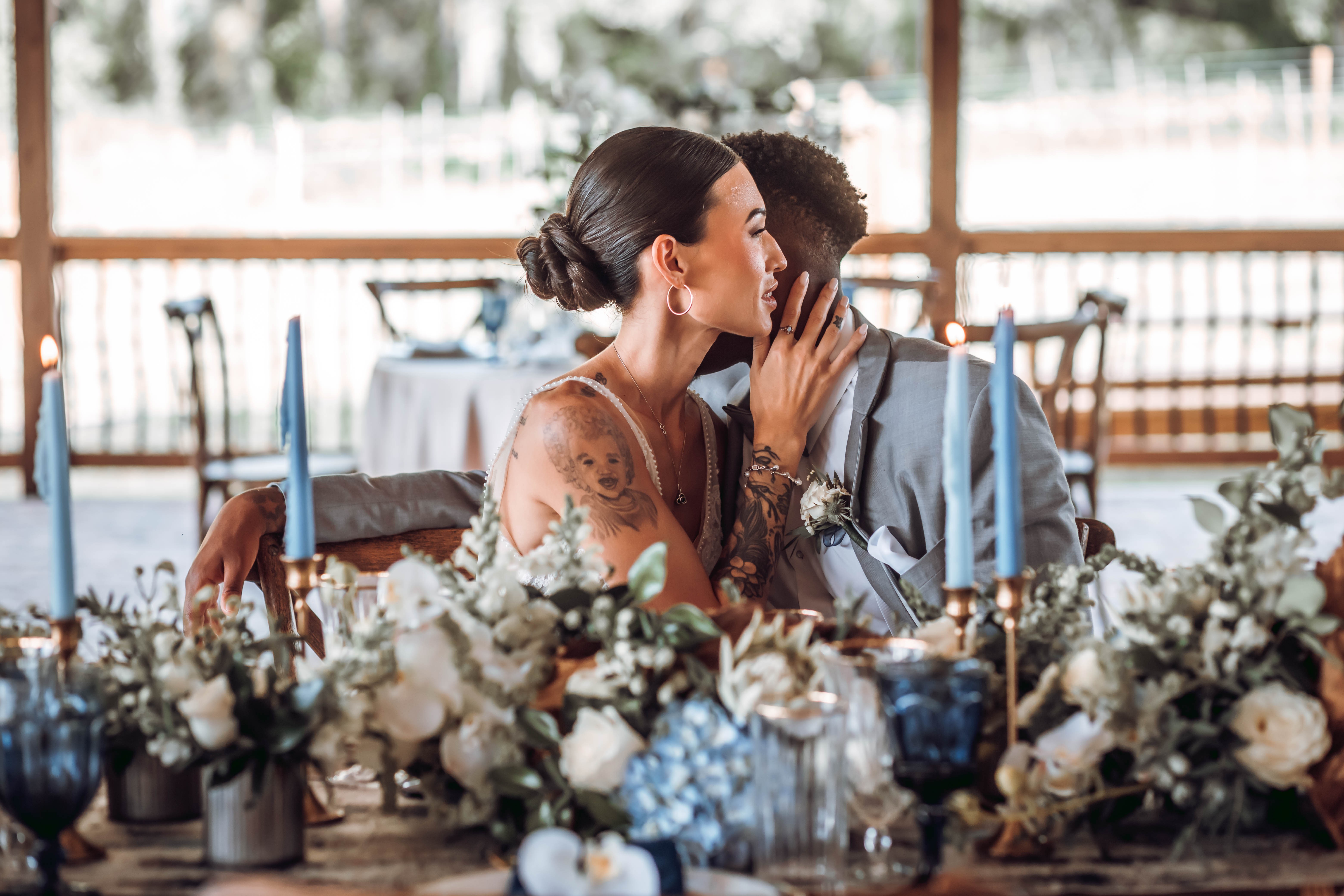 Two people sharing a kiss at a decorated table with flowers and candles, outdoors with a wooden railing and trees in background.
