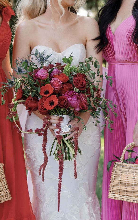 Woman in white dress holding a large bouquet of pink, red, and purple flowers with greenery, surrounded by people in colorful dresses.