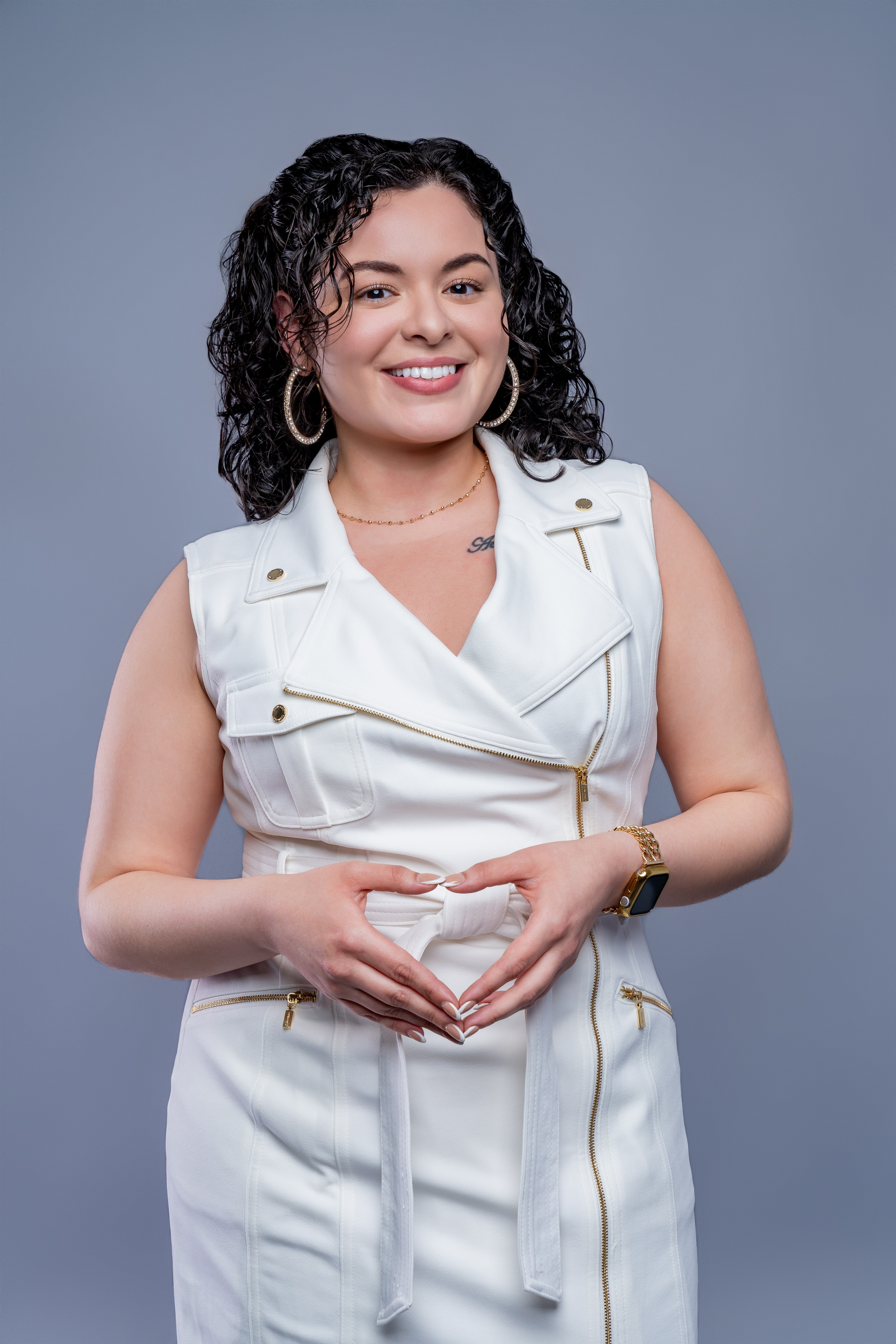 Young woman with curly hair smiling, wearing a white sleeveless outfit, standing against a gray background.