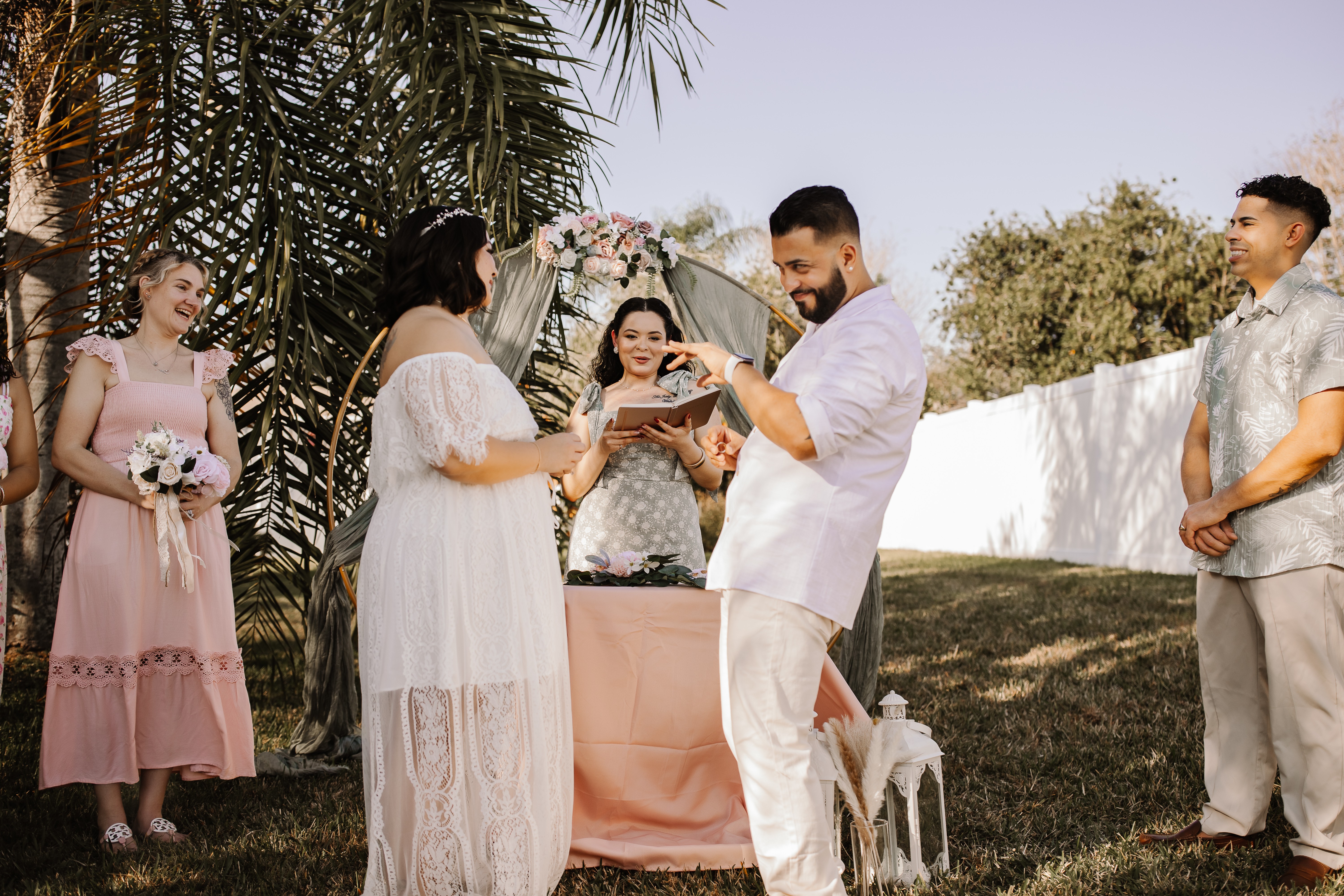 Group of people outdoors, with a woman in white exchanging rings with a man in white, others watching, trees in background