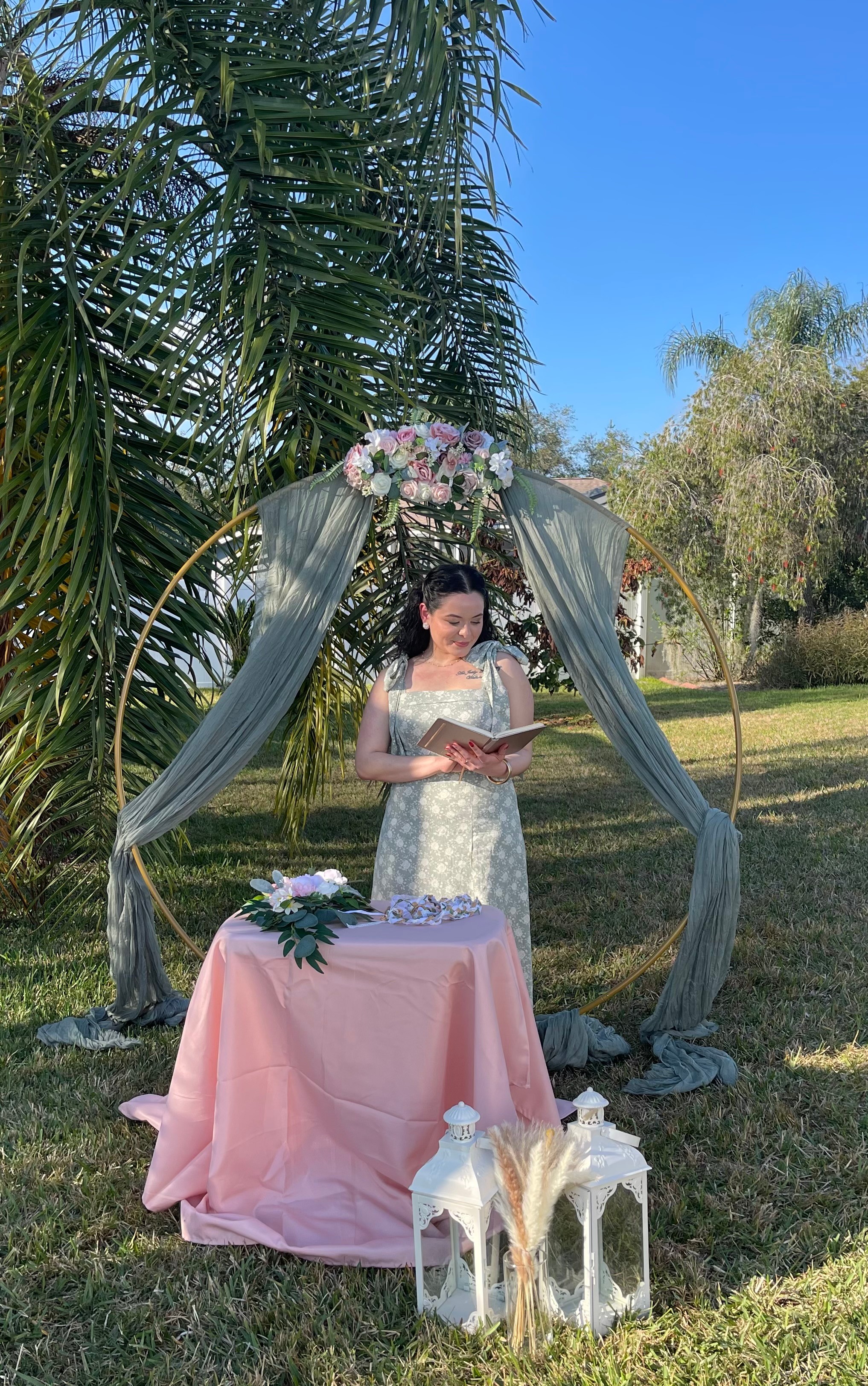 Woman in a white dress standing under a decorated arch outdoors, with a pink table and lanterns nearby, in a garden setting.