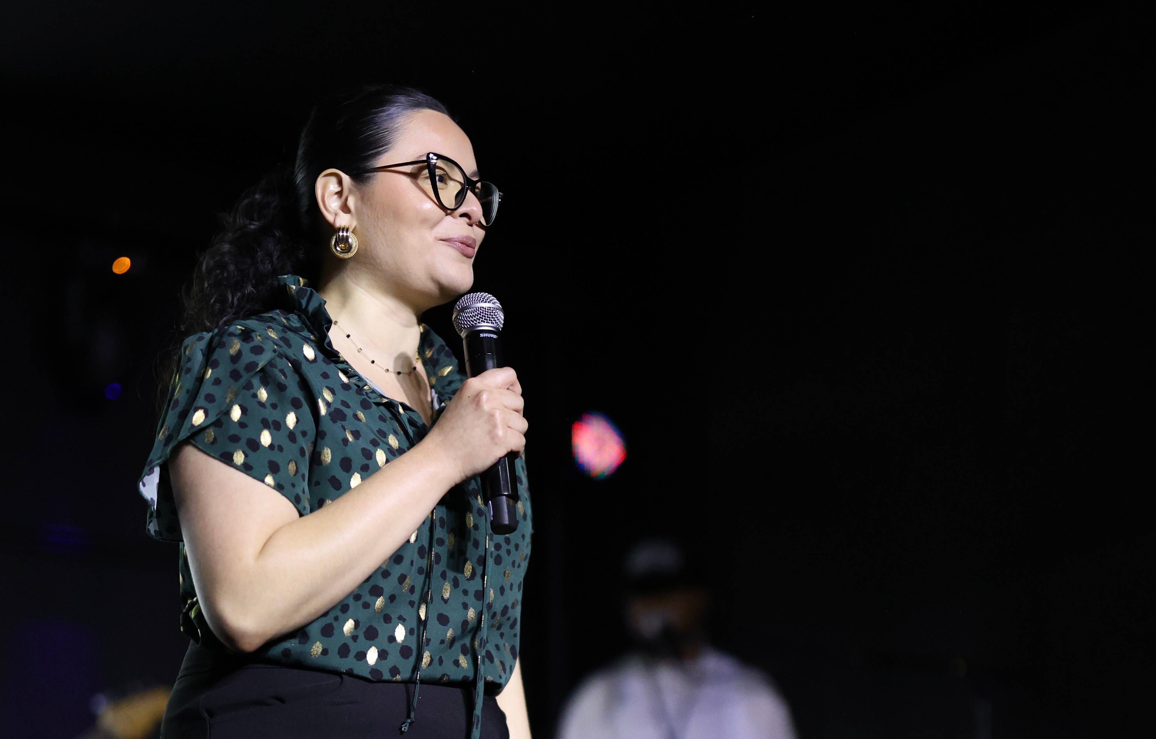 Woman speaking into a microphone, wearing glasses and a polka dot blouse, on stage with a dark background.