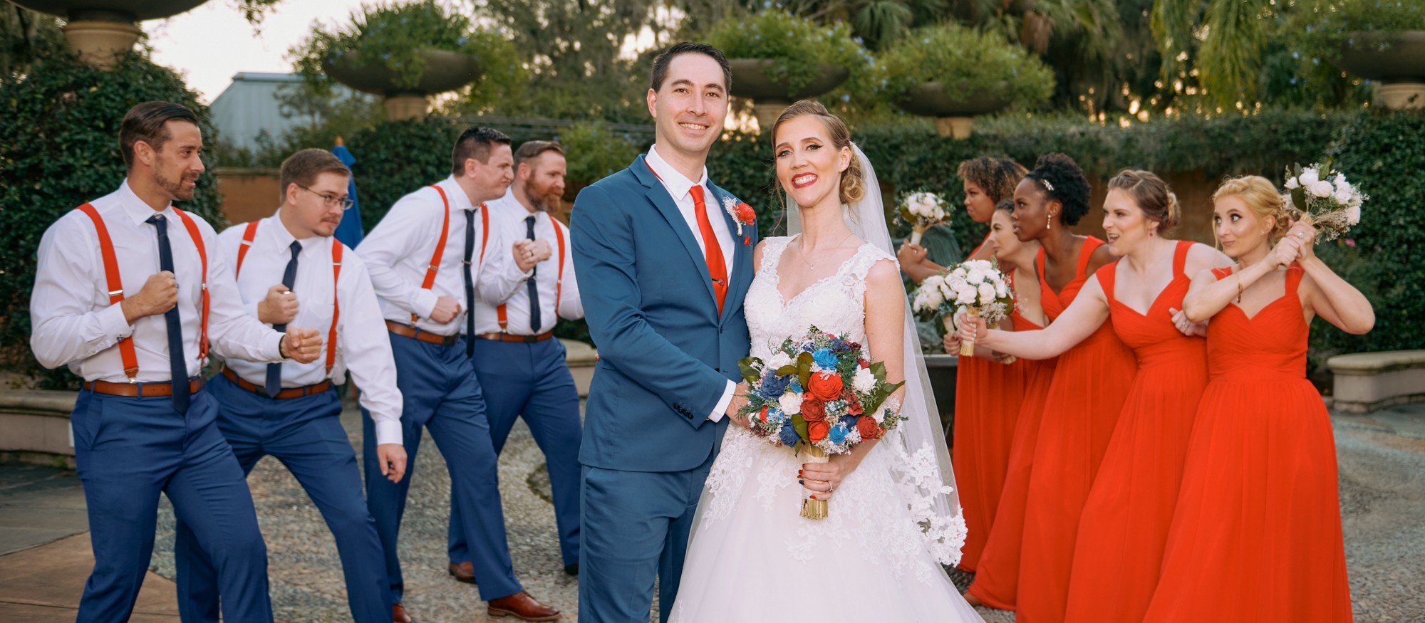 Wedding couple with bridesmaids and groomsmen outdoors, smiling, holding flowers, in formal attire.