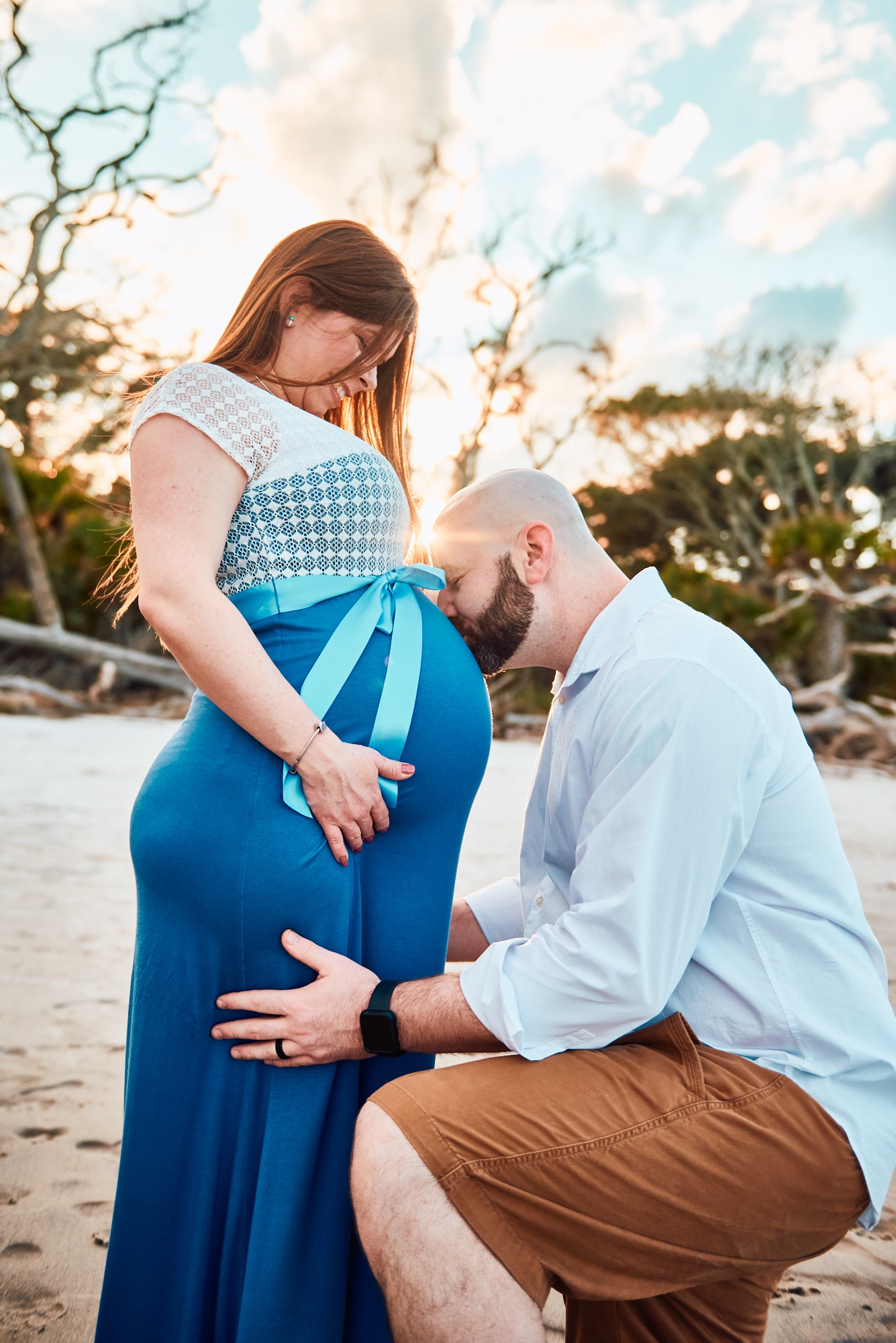 Pregnant woman in a blue dress with a man kneeling and kissing her belly outdoors, trees and sky in background.