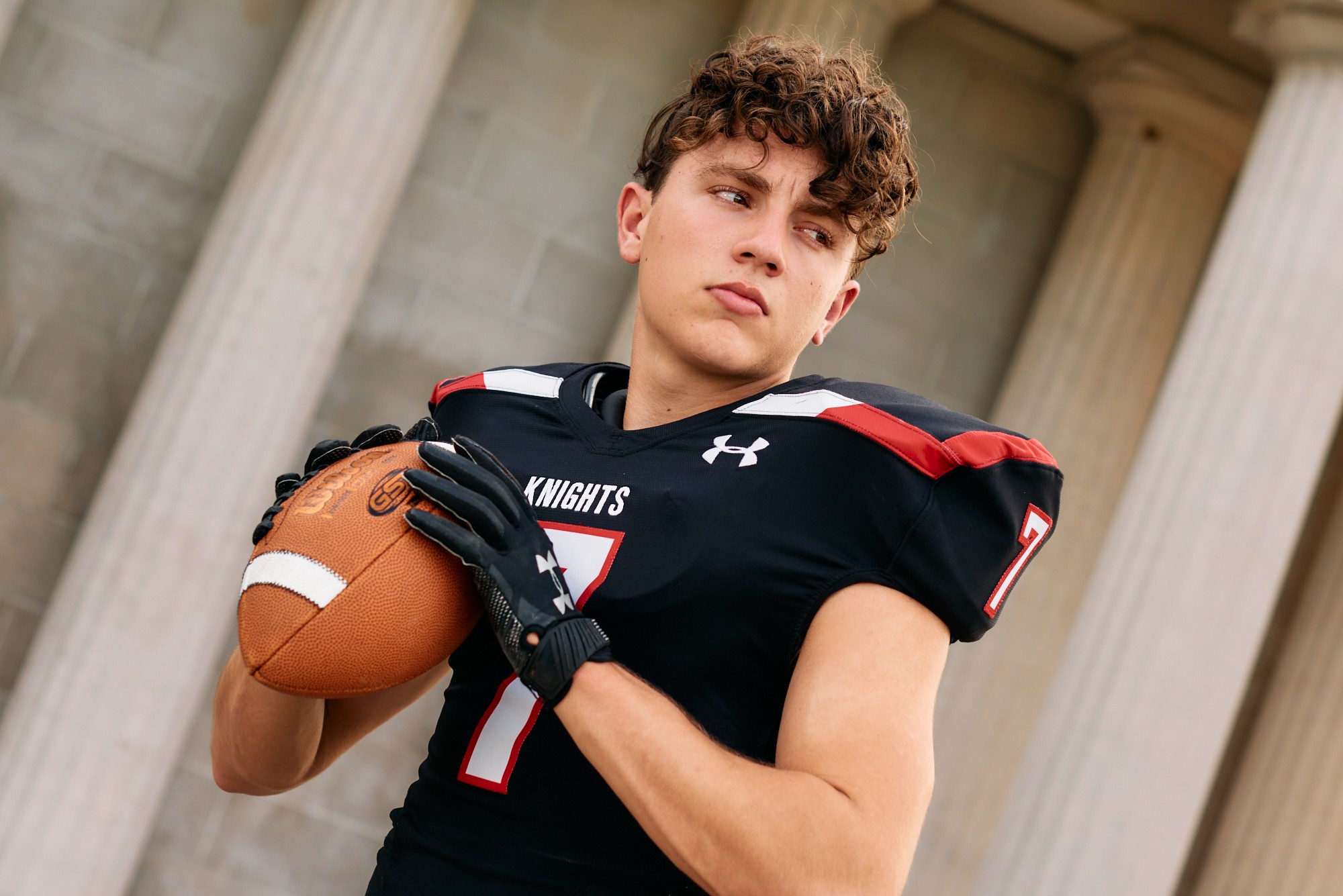 Young male football player in uniform holding a football, standing outdoors with columns in the background.