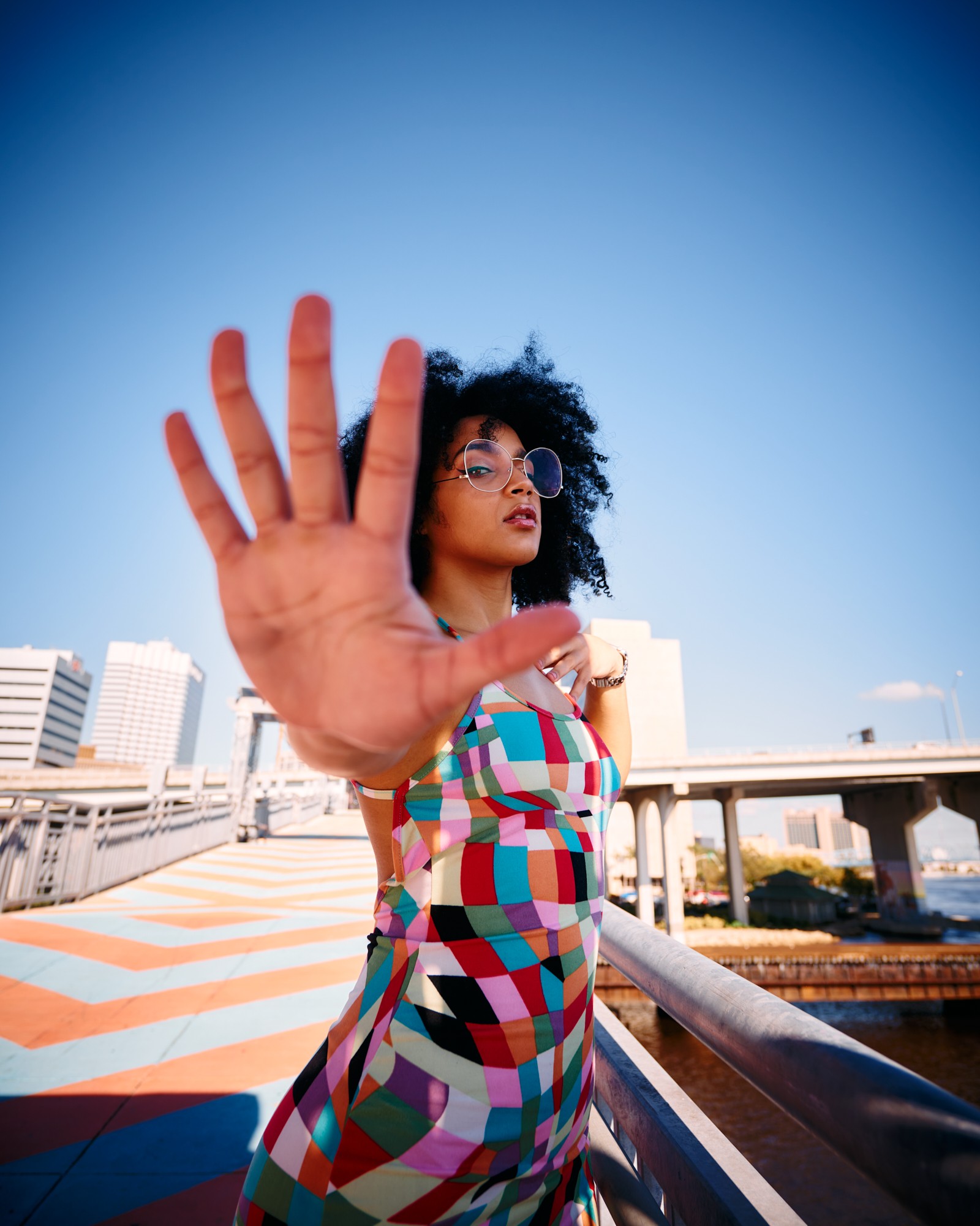 Woman with curly hair and glasses extends hand toward camera on city bridge, blue sky background.