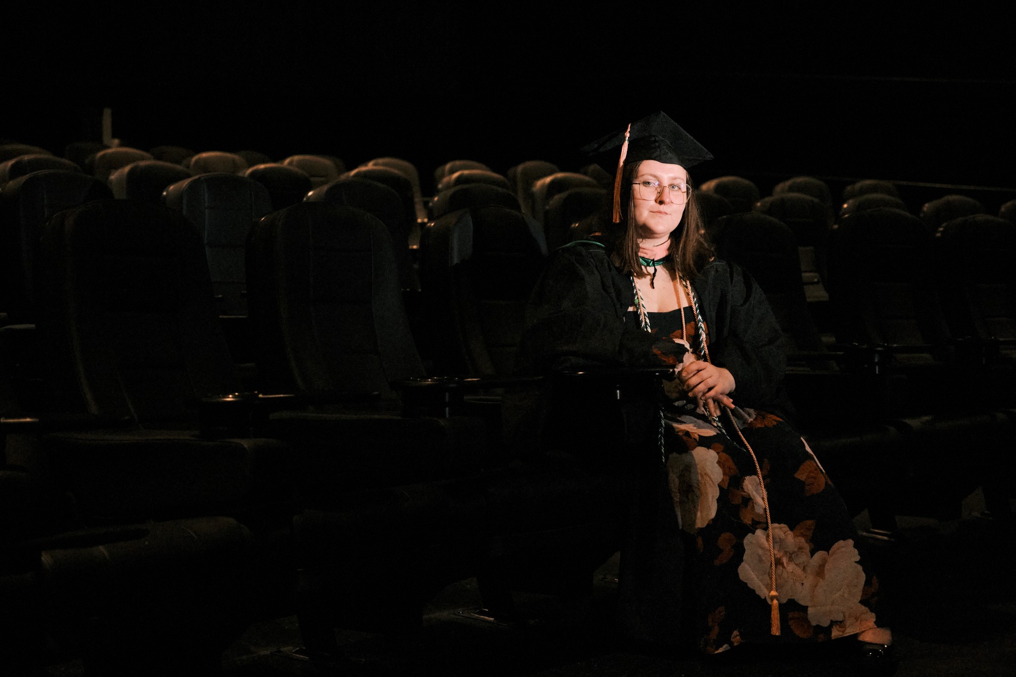 Woman in graduation cap and gown holding a diploma, seated in a dark auditorium with empty chairs.