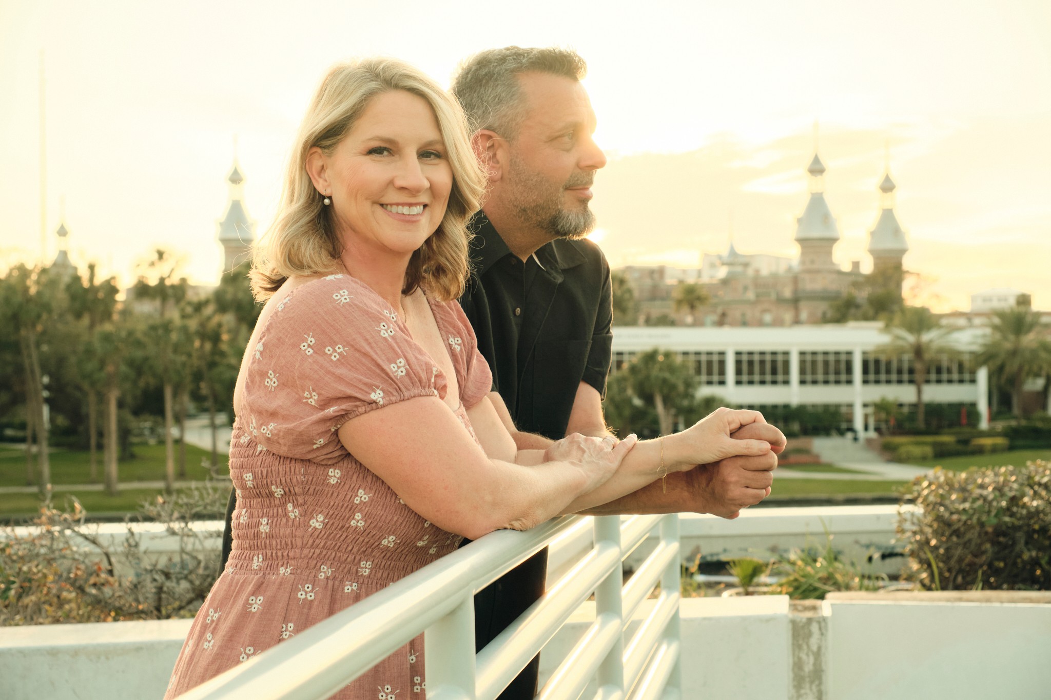 A smiling woman and a man leaning on a white railing outdoors with a cityscape and church spires in the background.