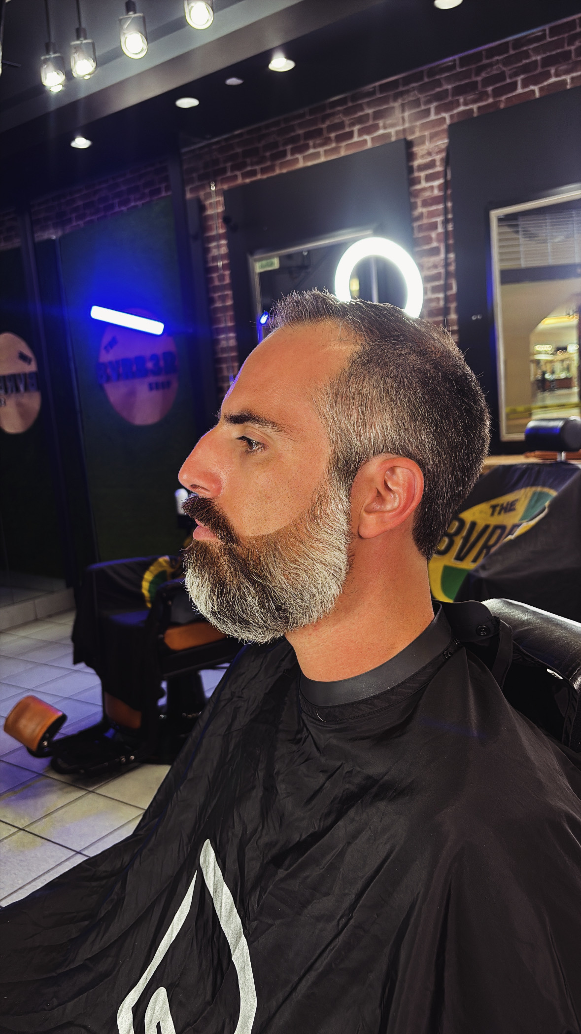 Side profile of a man with a beard and short hair, sitting indoors with a brick wall background.