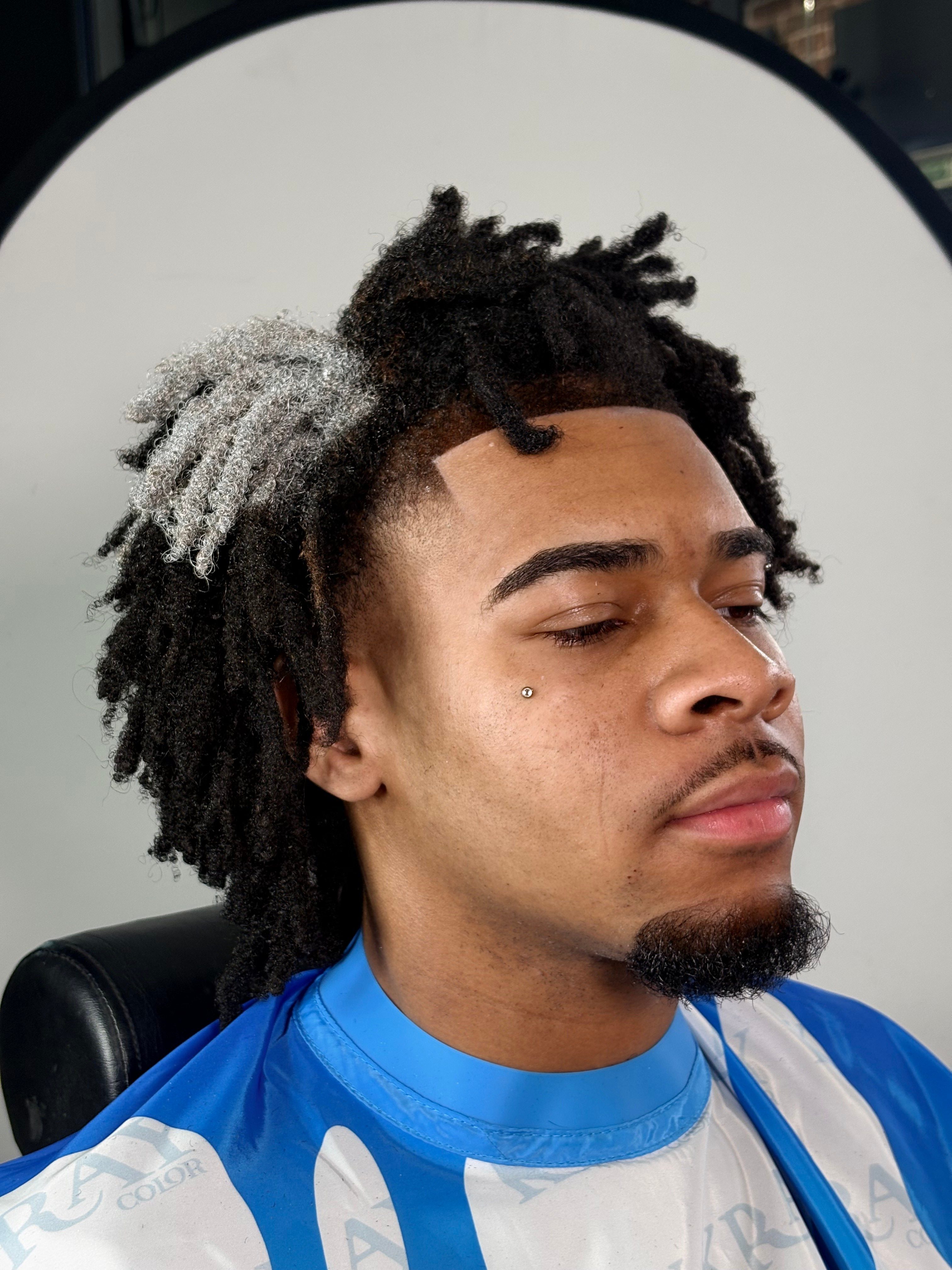 Young man with dreadlocks and facial hair, lying down, wearing a blue and white sports jersey, smiling slightly.